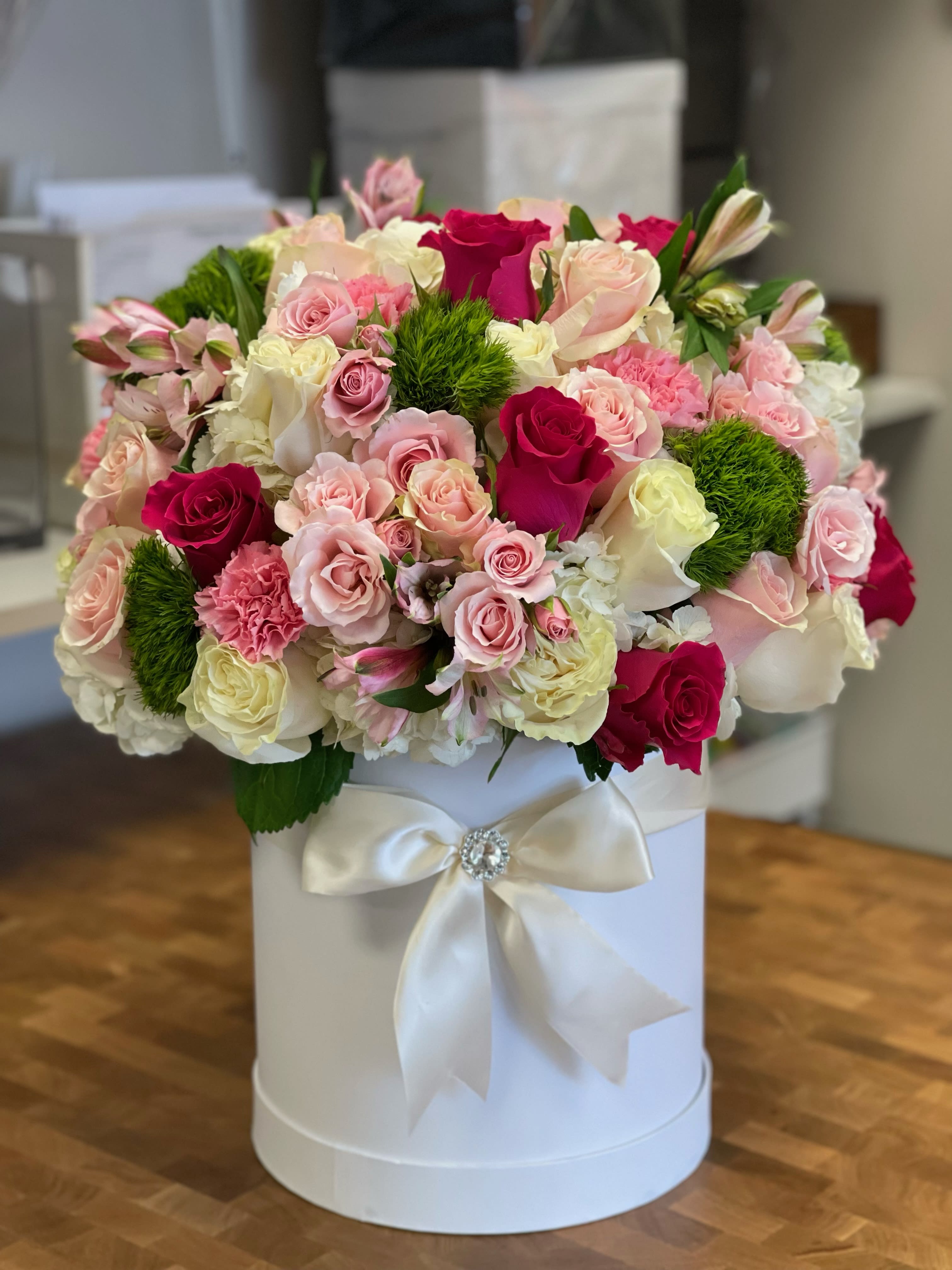 Round arrangement of pink and white roses with green accents in a white hatbox with satin bow