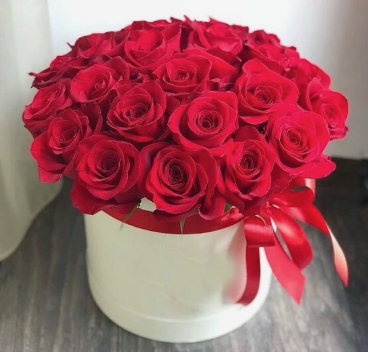 Round box of tightly arranged red roses with a red ribbon bow on a white container