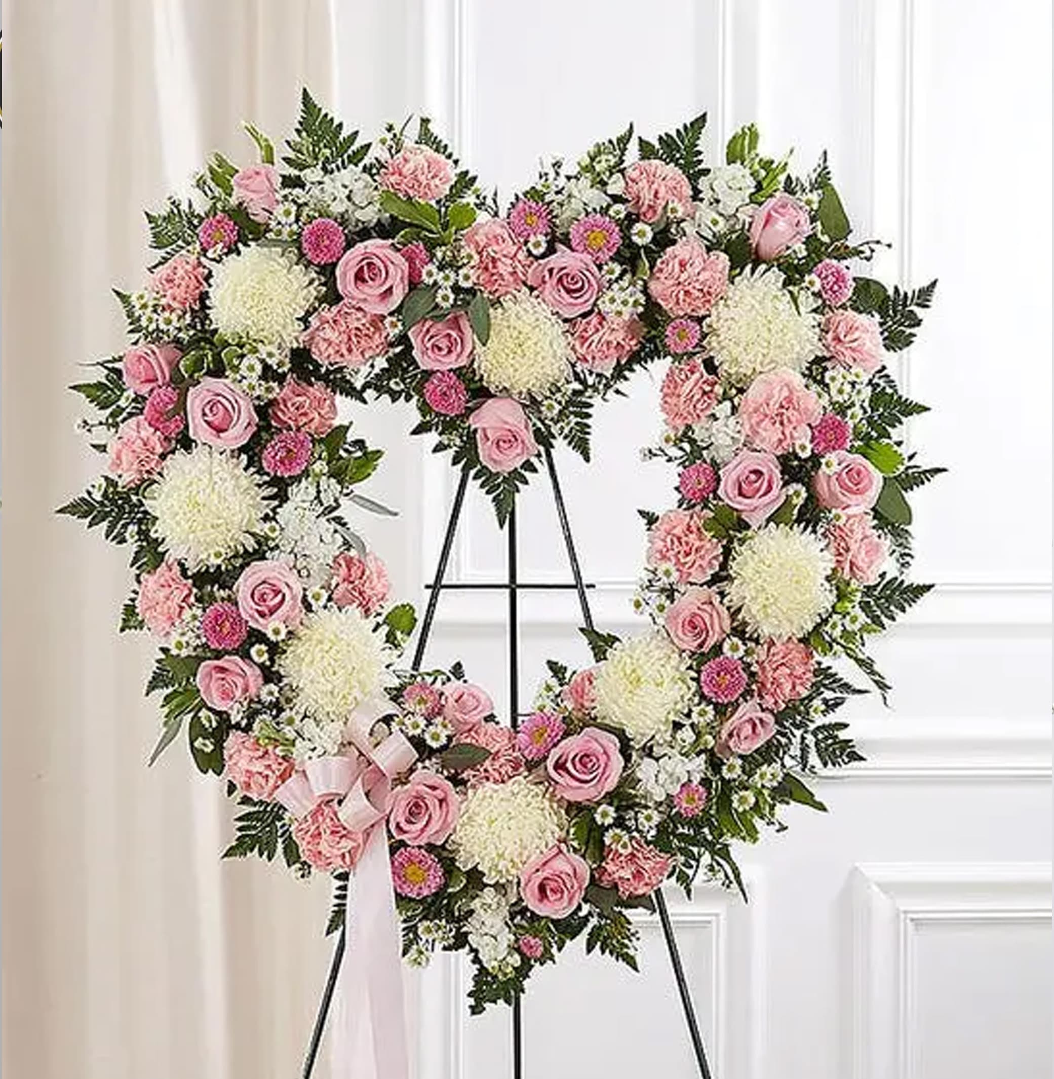 Heart-shaped standing wreath of pink roses, carnations, and white mums on a metal easel with a pink ribbon bow