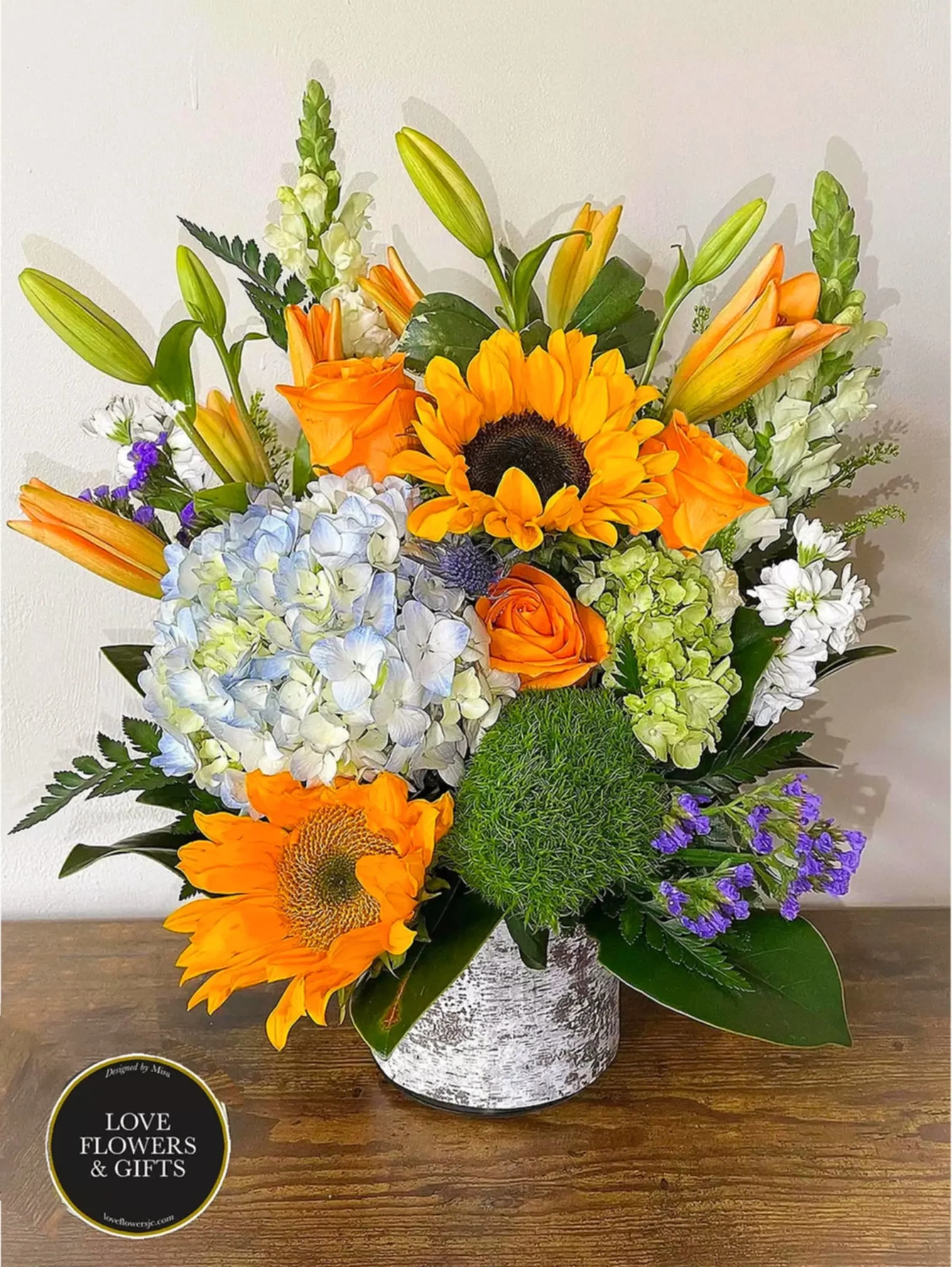 Bright arrangement of sunflowers, orange roses, lilies and hydrangeas in a white cylinder vase