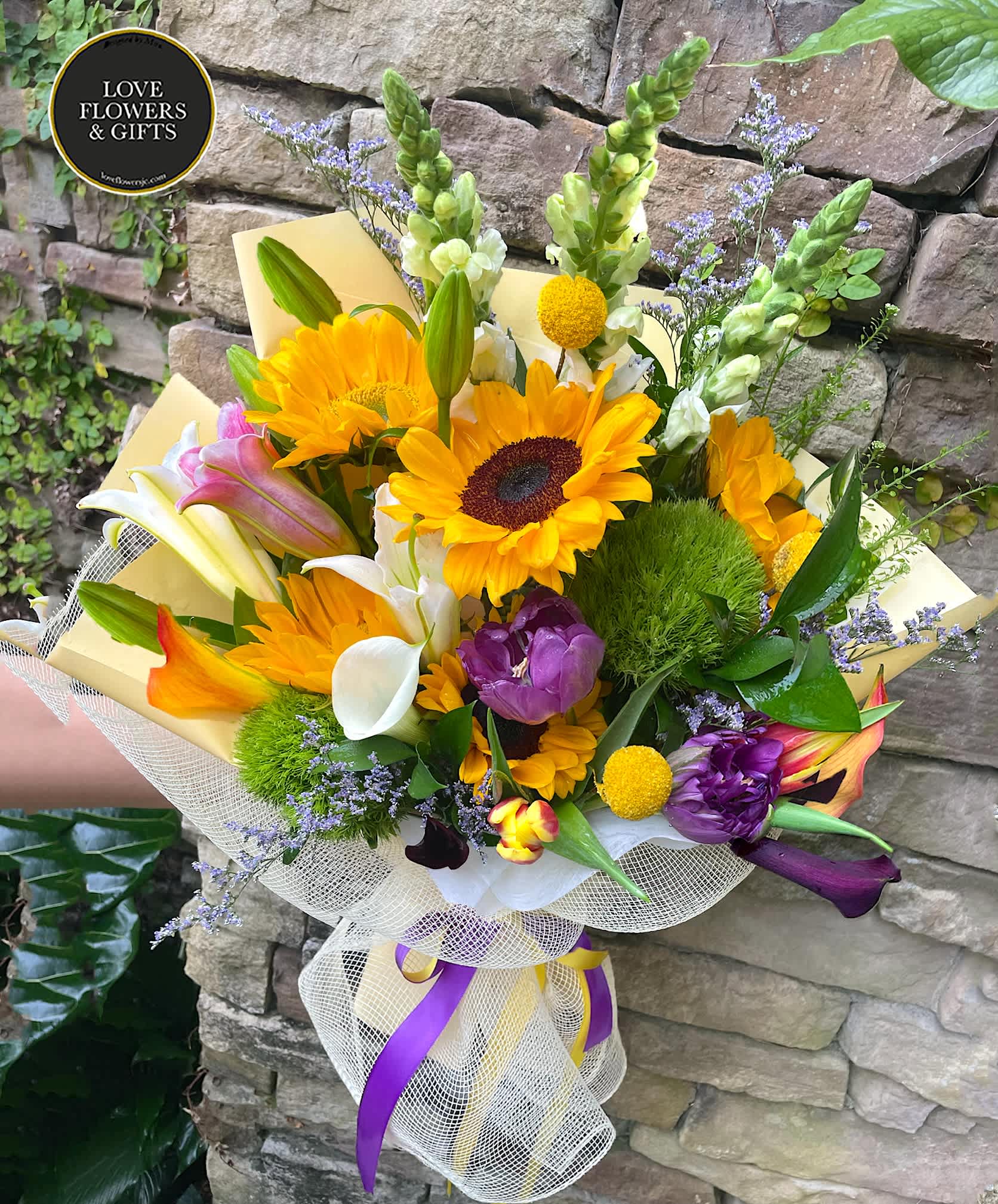 Bright mixed bouquet with sunflowers, lilies, snapdragons and purple blooms wrapped in mesh with ribbons.