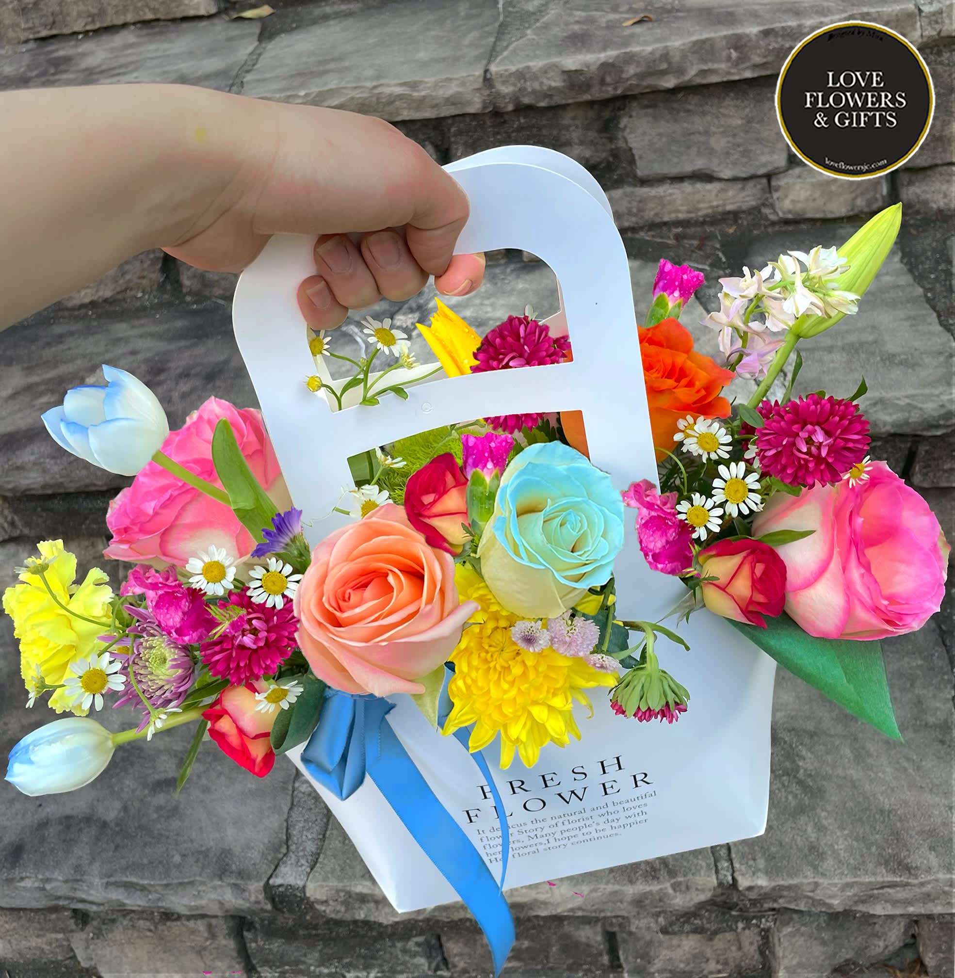 Colorful mixed bouquet of roses, daisies, and mums in a white carry bag with handle