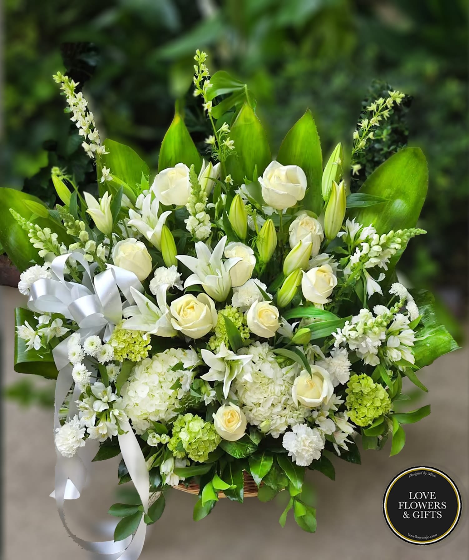 Large white and green sympathy basket with roses, lilies, hydrangeas, and a white ribbon bow.