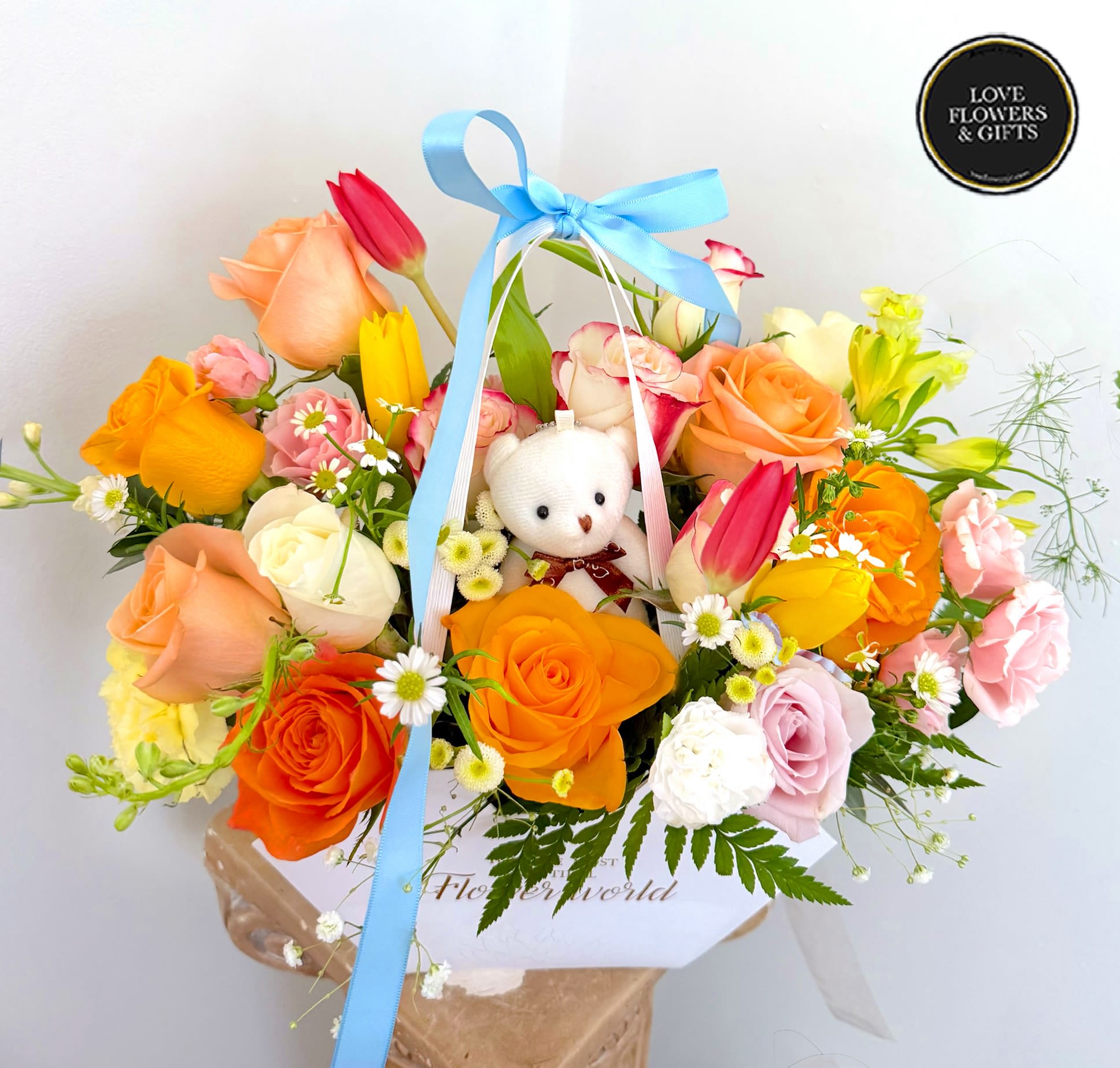 Bright mixed arrangement of roses and tulips with a small white teddy bear in a white box.