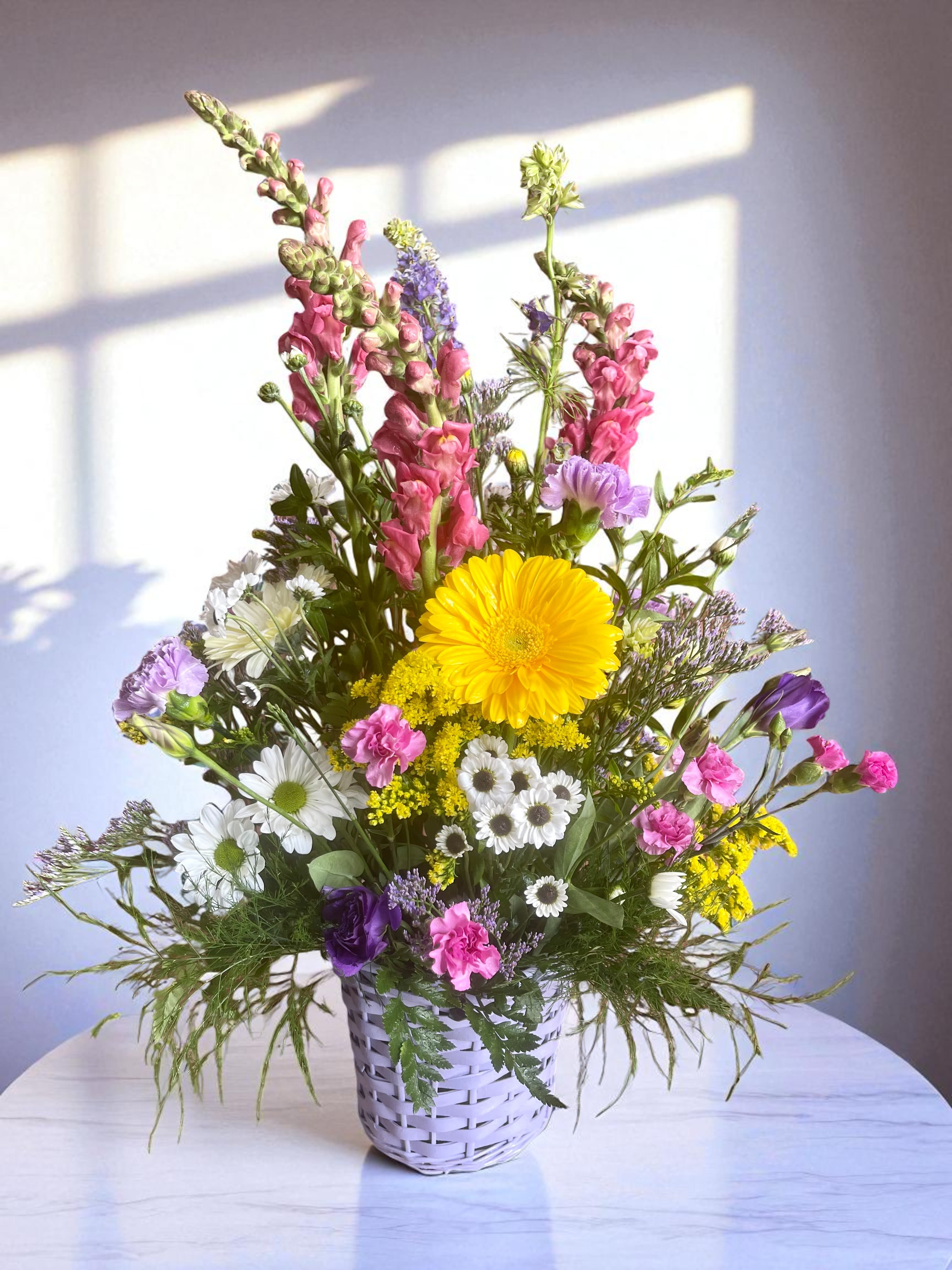 Mixed bouquet in a white woven basket with yellow, pink, purple, and white flowers