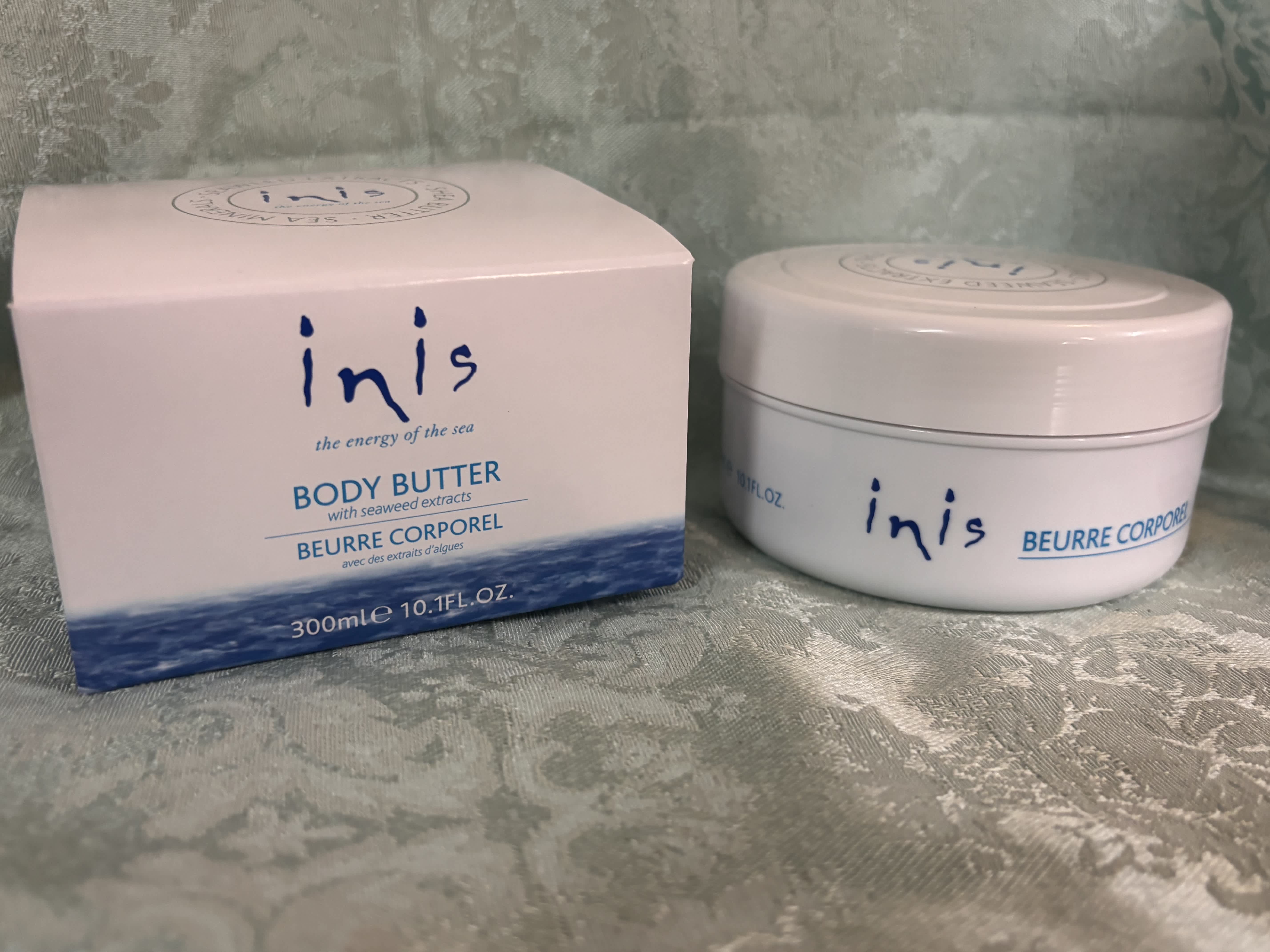 Box and jar of Inis body butter on a patterned fabric surface