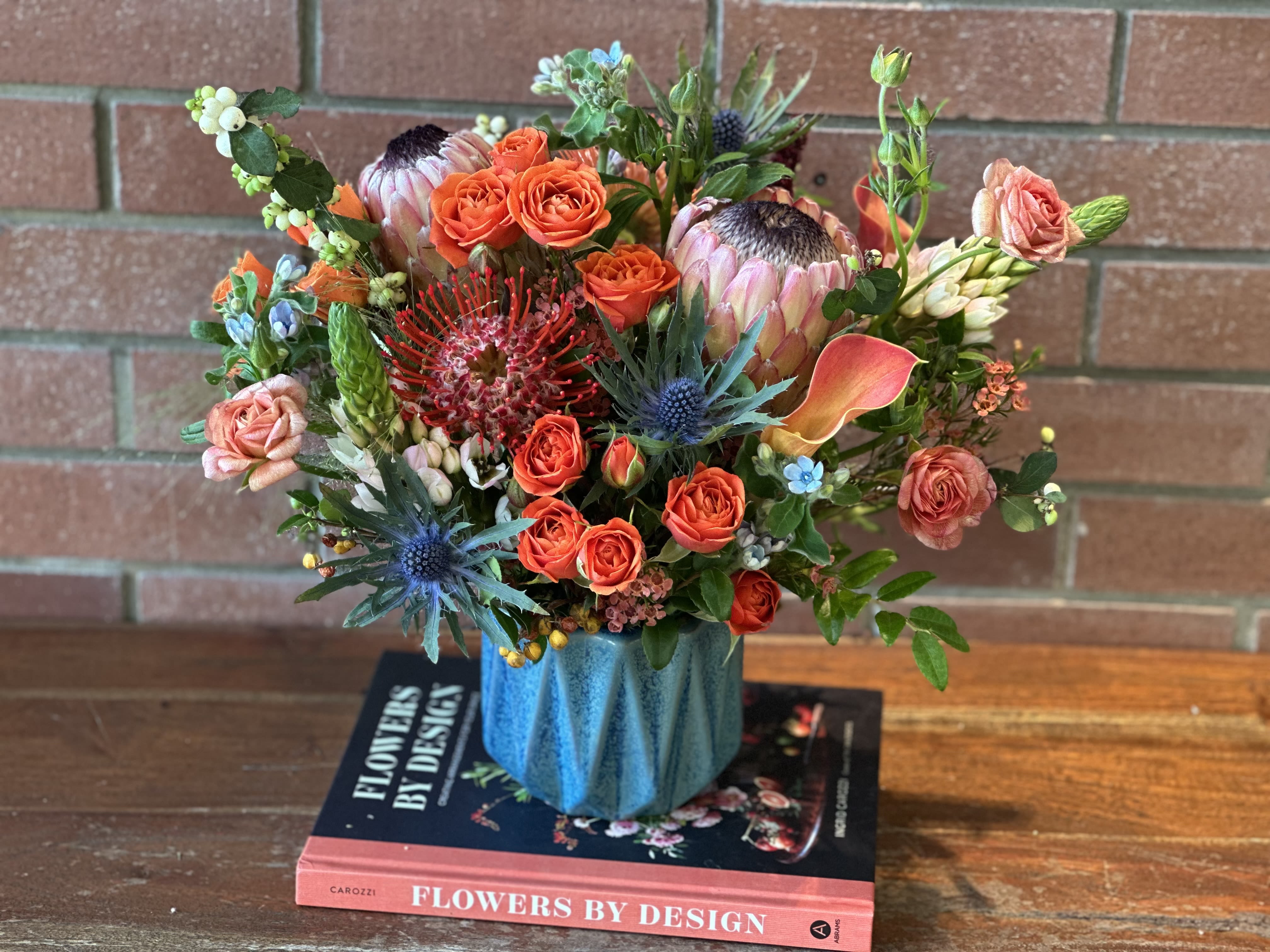 Colorful arrangement with orange roses, protea, and calla lilies in a blue vase on books