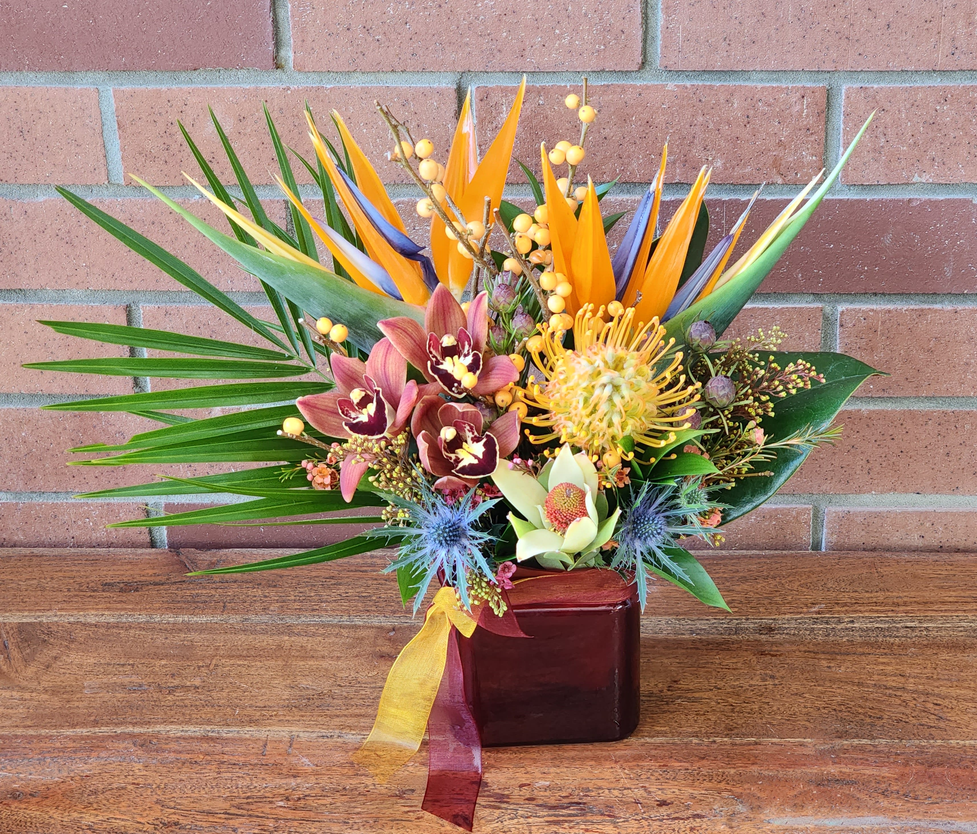 Tropical arrangement with birds of paradise, orchids, and protea in a red glass cube vase with ribbon