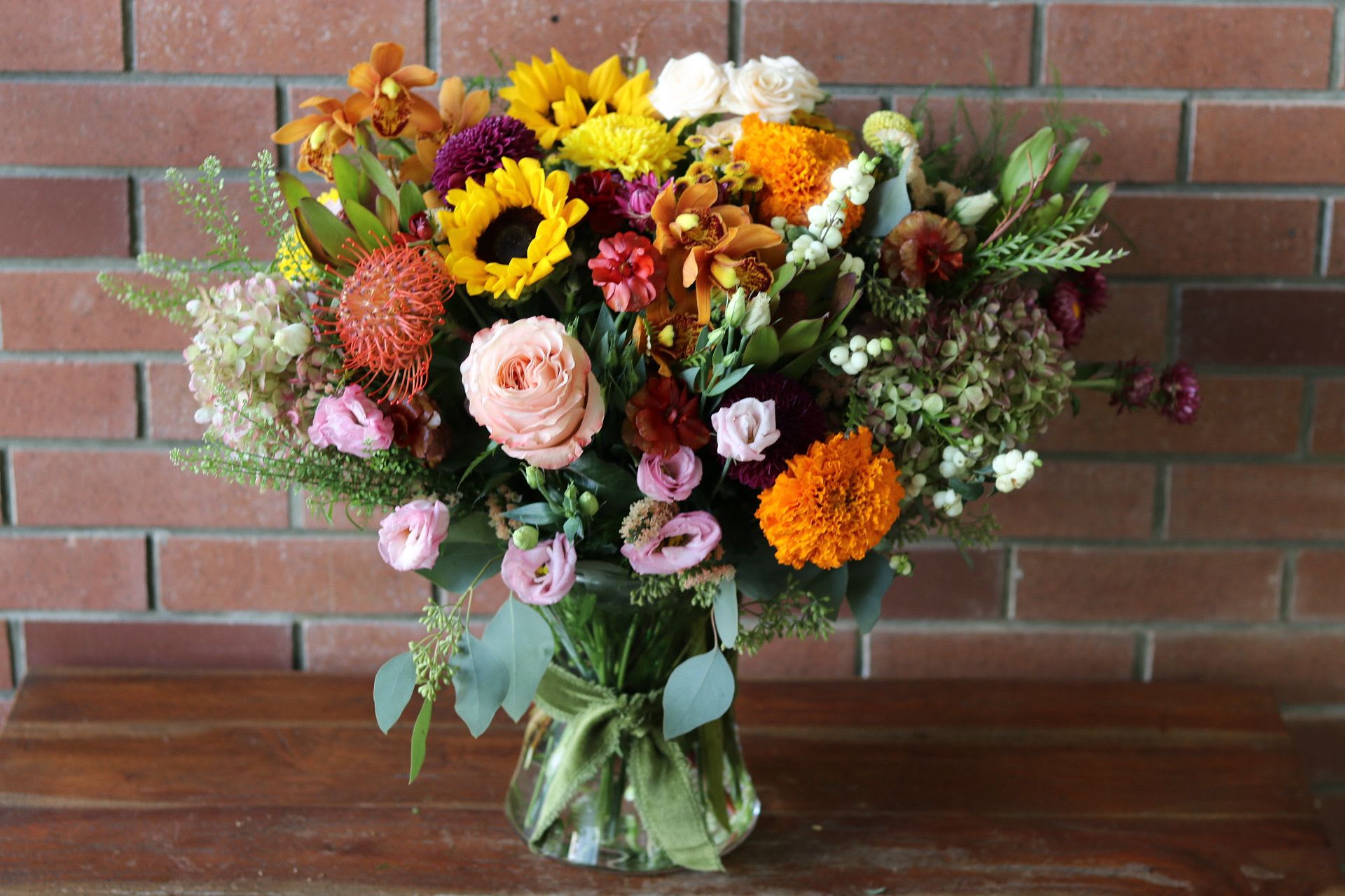 Large Farm Fresh Fall Bouquet  - The Farm Fresh Fall Bouquet by Venetian Flowers is a stunning celebration of autumn's vibrant colors and textures. This arrangement features an exquisite selection of seasonal blooms, each chosen for their freshness and longevity.   At the heart of the bouquet are the striking ball mums and unique pin cushion proteas, adding a bold and dynamic visual element. The delicate lisianthus and bright orange orchids bring warmth and elegance, while the charming strawflowers and a variety of roses, including garden roses, introduce layers of softness and romance.   All of these beautiful flowers are artfully arranged in a clear vase, allowing the vibrant colors to shine through. The bouquet is further enhanced by lovely fringe ribbons that add a touch of whimsy and charm, making it a perfect centerpiece for any fall gathering or a thoughtful gift to brighten someone's day.
