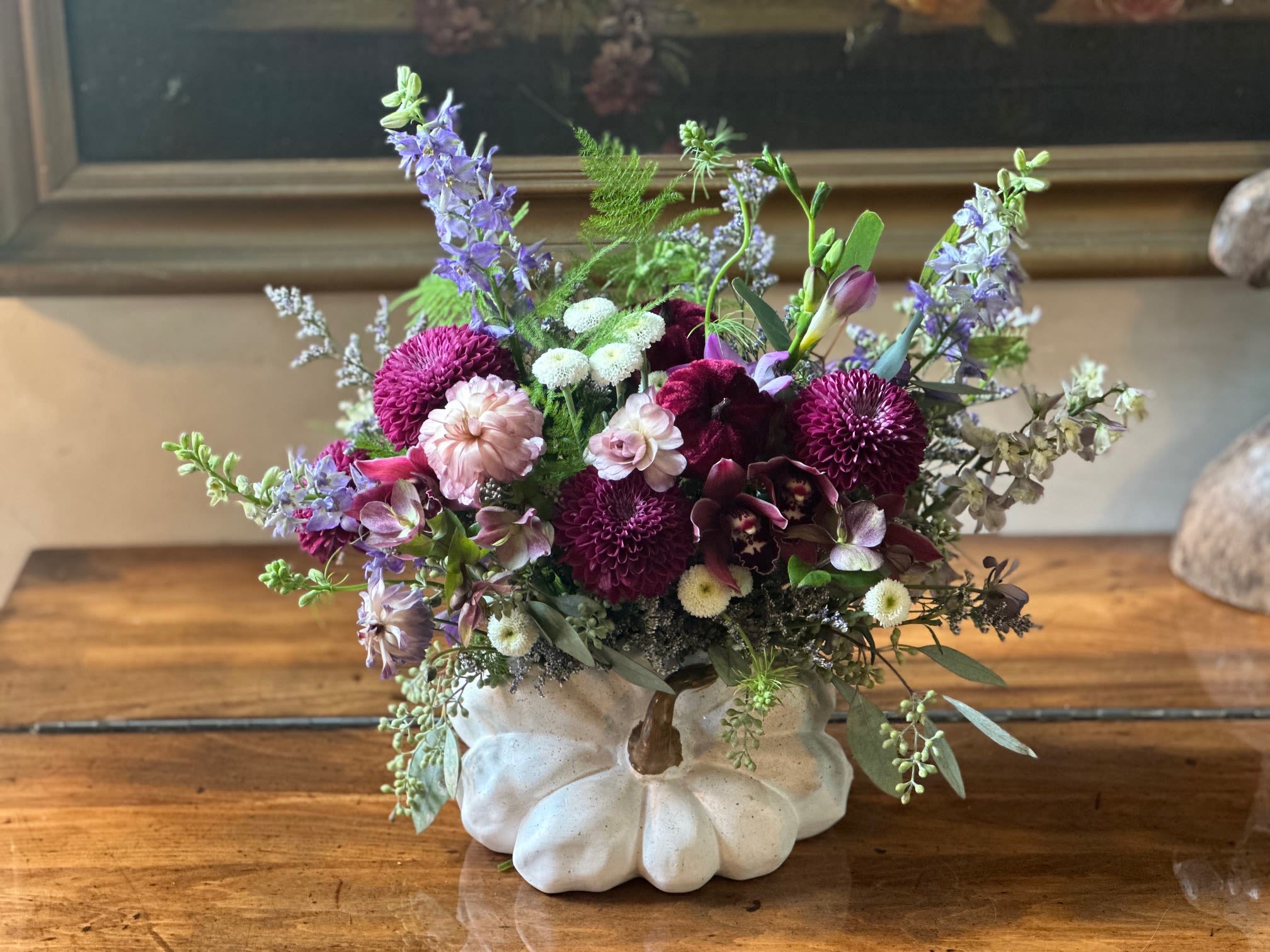 Autumn Enchantment Pumpkin - Celebrate the rich beauty of fall with our Autumn Enchantment Arrangement, artfully designed in a charming white pumpkin vase. This stunning creation features purple larkspur reaching skyward, luxurious purple ball chrysanthemums, delicate white button mums, and exquisite mini cymbidium orchids in rich fall hues. Soft ranunculus blooms add romantic texture, while lush seasonal greenery completes this captivating display. Perfect for bringing the warmth and elegance of autumn into any space.