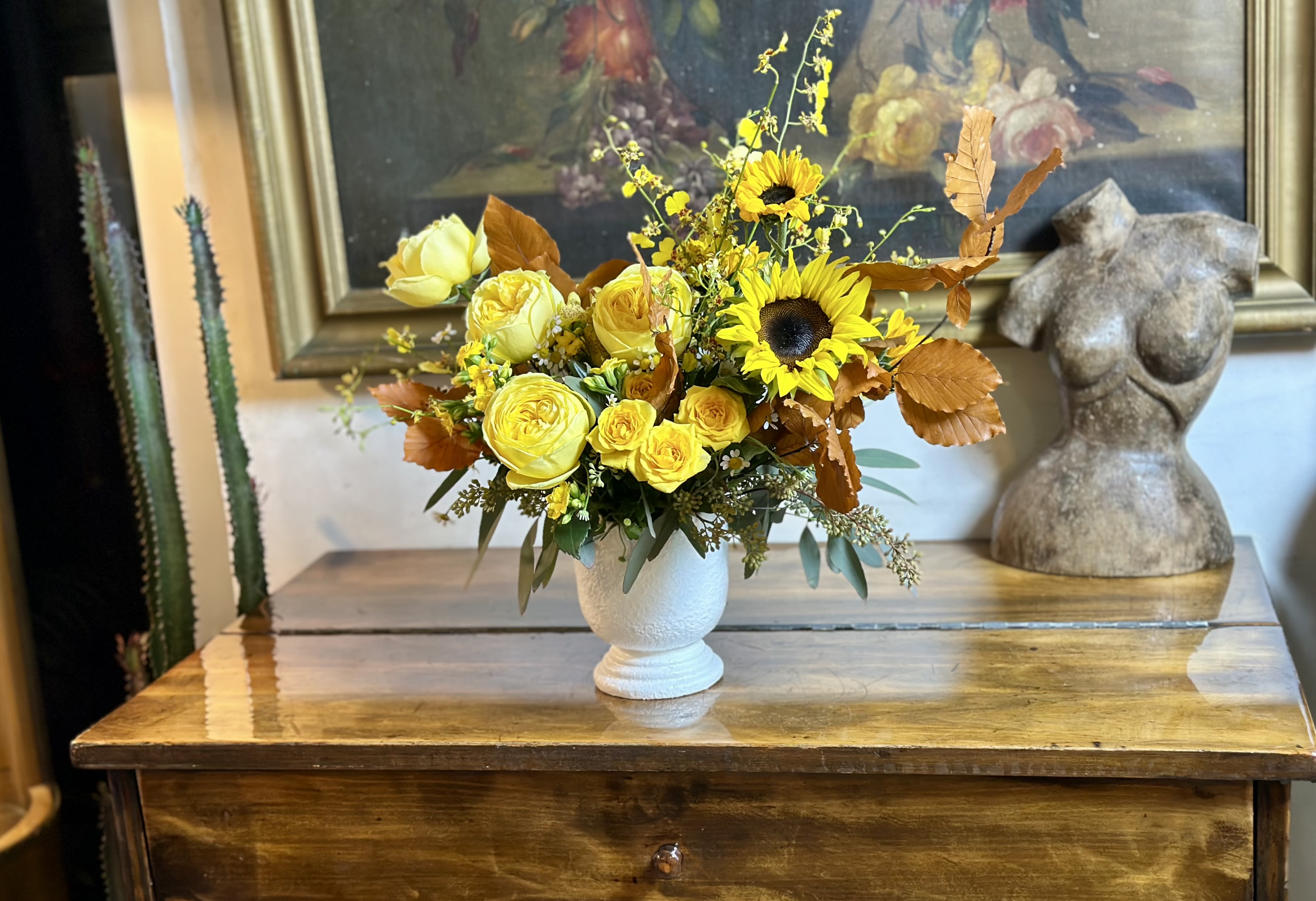 Autumn Sunshine  - Brighten your Thanksgiving celebration with the radiant warmth of this golden masterpiece. “Autumn Sunshine” captures the luminous beauty of fall with its brilliant display of fresh sunflowers, luxurious garden roses, and cheerful yellow spray roses that glow like autumn’s golden light. Delicate dancing oncidium orchids add graceful movement and an elegant surprise, while rich copper oak leaves bring the authentic textures of the season indoors. Eucalyptus and seasonal fillers provide lush depth, creating a full, abundant arrangement that embodies the spirit of harvest and gratitude. Displayed in an elegant textured white ceramic vase, this arrangement brings instant warmth and joy to your holiday table. Perfect for those who love the classic beauty of fall but want to keep things bright and uplifting, “Autumn Sunshine” is a cheerful reminder of all the reasons we gather to give thanks. Features: 	•	Fresh sunflowers 	•	Oncidium orchids 	•	Premium garden roses 	•	Yellow spray roses 	•	Copper fall oak leaves 	•	Eucalyptus 	•	Seasonal blooms and premium fillers 	•	Textured ceramic vase