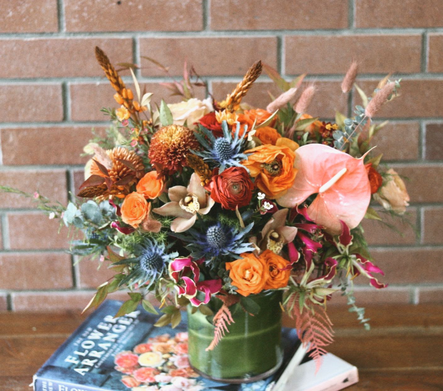 Autumn Luxe  - Arrangement Name: "Citrus Luxe"   Description:   Immerse yourself in the vibrant hues of fall with "Autumn Luxe," a stunning modern bouquet crafted by Venetian Flowers. This exquisite arrangement features the exotic beauty of Gloriosa lilies, their fiery petals dancing alongside the cheerful orange ranunculus and the sleek, tropical anthurium. Accentuating the warmth of the arrangement are the delicate cymbidium orchids and playful orange spray roses, bringing a touch of elegance and joy.  The bouquet is beautifully complemented by the striking blue thistle, which introduces a bold contrast, while the playful orange ball mums add texture and whimsy. Delicate Star of Bethlehem flowers peek through, adding a touch of ethereal charm, while seasonal fillers and lush greenery provide a rich backdrop that enhances the overall composition.  All of this is artfully arranged in a sleek modern cylinder vase, elegantly lined with a fresh green leaf that adds an extra layer of sophistication. "Autumn Luxe" is perfect for any occasion, bringing the warmth and vibrancy of the fall season into your space. Let this bouquet be a stunning centerpiece that captures the beauty of nature's palette.