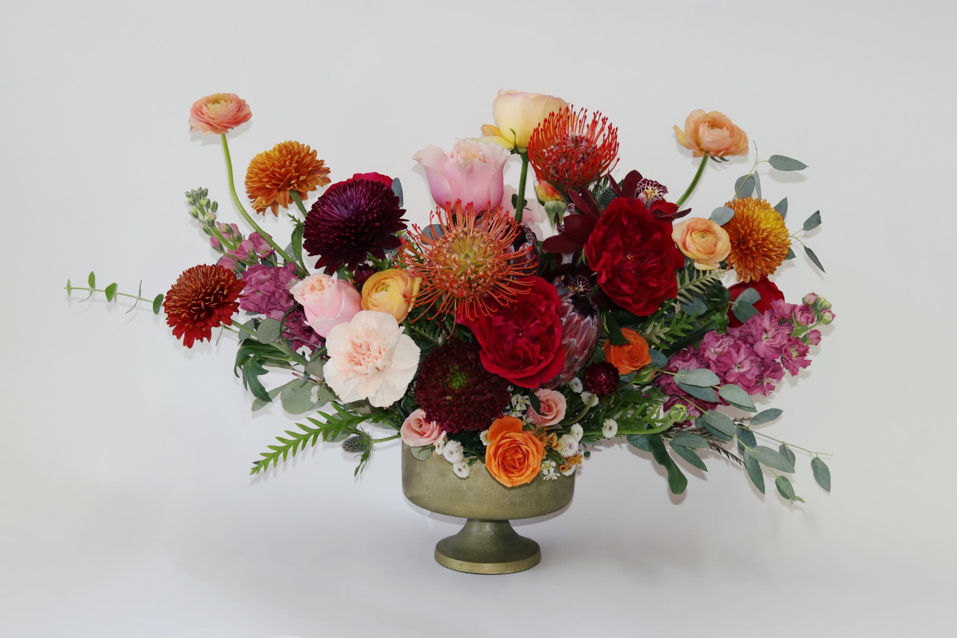 Autumn’s Embrace - **Name:** "Autumn's Embrace"  **Description:**   "Autumn's Embrace" is a stunning autumn arrangement from Venetian Flowers' 2024/2025 collection, designed in a modern gold compote vase. This bouquet beautifully encapsulates the essence of fall with its rich palette of orange, burgundy, and peach blooms.  Lush ranunculus and garden roses add a romantic touch, while the unique pin cushion proteas and burgundy mini cymbidium orchids serve as striking focal points with their intriguing textures. Vibrant mums and delicate spray roses, stock, enhance the bouquet's softness and depth, while thistle introduces a playful contrast.   Completed with an assortment of seasonal fillers and greenery, "Autumn's Embrace" radiates warmth and charm, making it an ideal centerpiece for fall gatherings or a thoughtful gift that captures the spirit of the season.gundy 
