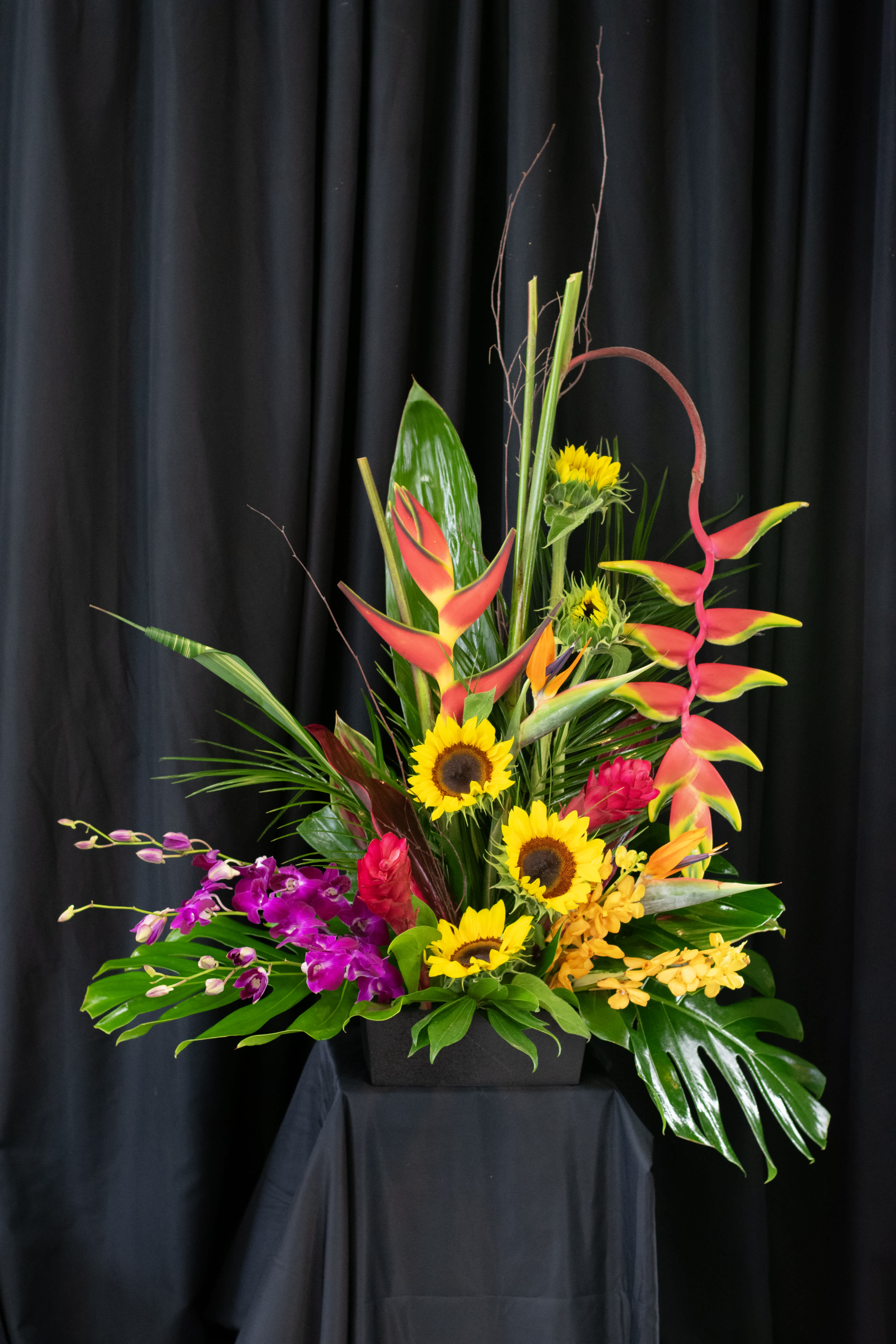 Tropical arrangement with sunflowers, orchids, and red heliconia in a black container