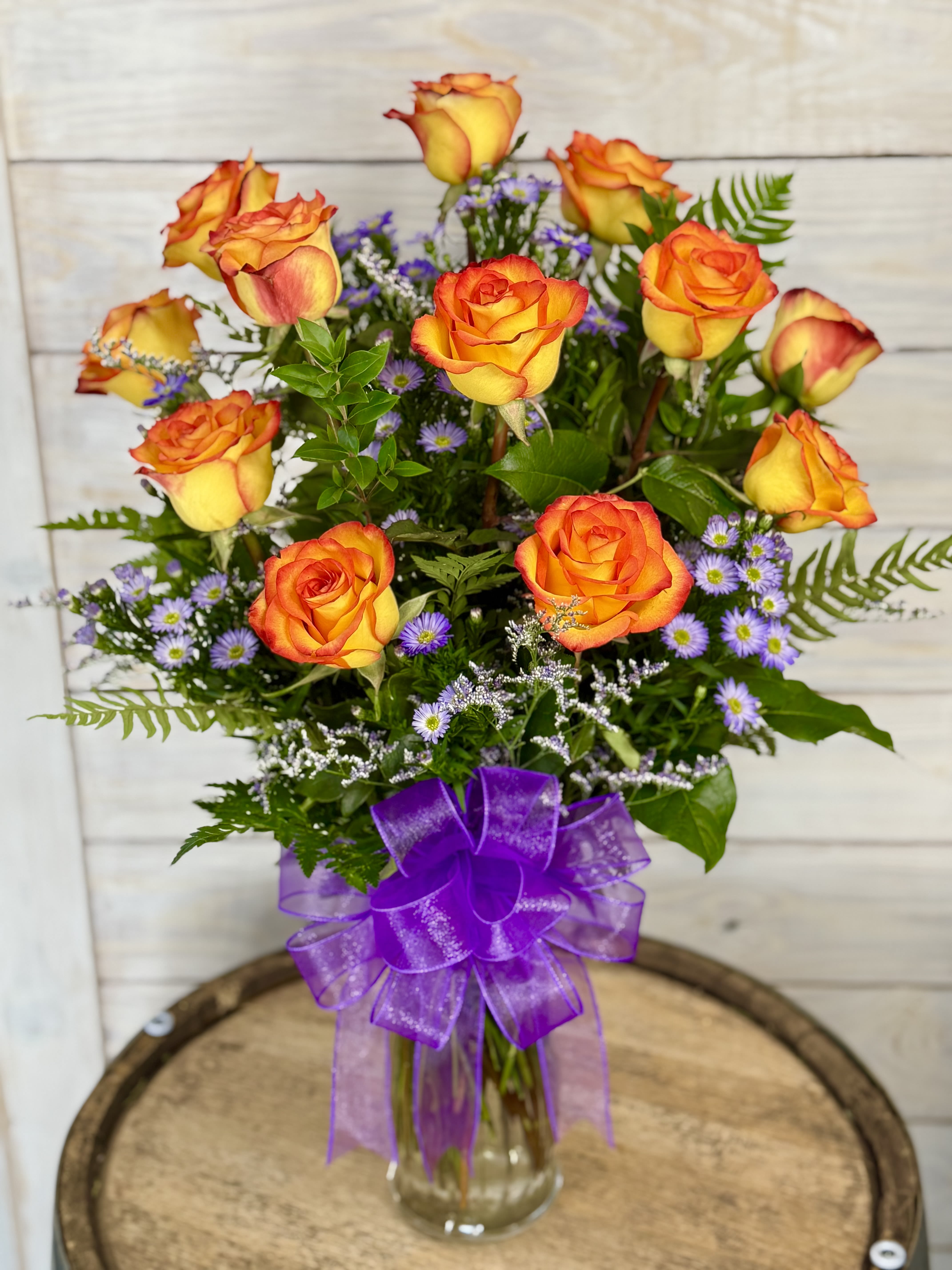 Orange-yellow roses in a glass vase with purple filler flowers and a purple ribbon