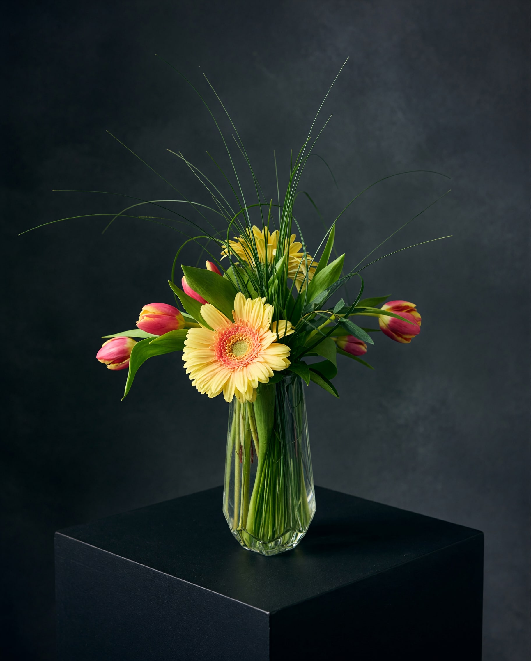 Sunlit Garden - Bright, graceful, and full of movement, Sunlit Garden brings the beauty of a fresh garden indoors. Sunny yellow gerbera daisies and delicate tulips are accented with lush greenery and airy grasses for a natural, modern look.  Designed in a clear glass vase, this arrangement makes a beautiful gift for birthdays, celebrations, congratulations, or simply letting someone know you’re thinking of them.    Flower varieties and colors may vary based on seasonal availability while maintaining the overall style, color palette, and value of the design.