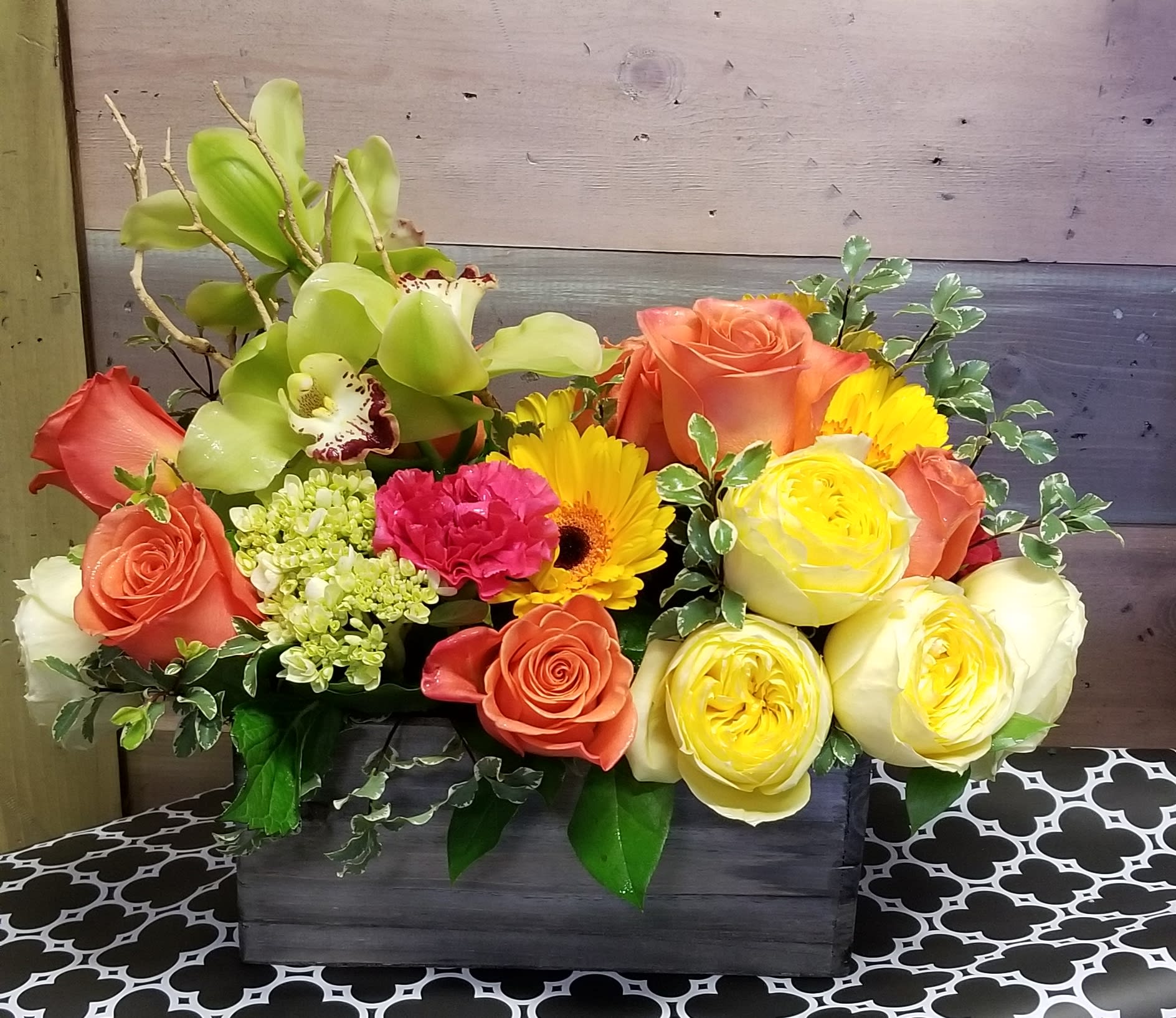 Low wood box arrangement with orange roses, yellow blooms, green orchids, and a pink carnation.