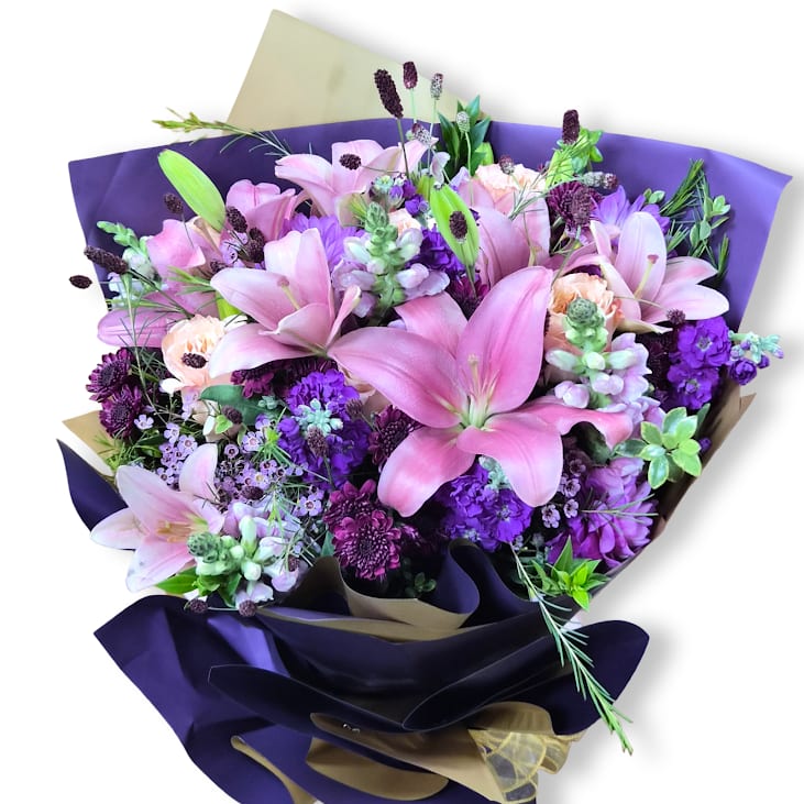 Purple Daydream Cello Wrap - A lush cascade of pink lilies and clustered purple stock makes a vivid first impression-perfect as a birthday bouquet, anniversary arrangement, or a cheerful "thank you" gift. The combination of saturated purples, soft pinks, and crisp whites creates a romantic yet upbeat mood that will make the person you're thinking of feel truly celebrated.  This flower arrangement pairs large, fragrant pink lilies with purple and lavender stock, chrysanthemums, and small airy accents for texture and depth. Greenery frames the blooms and a deep purple and tan decorative wrapping paper gives the bouquet a polished, gift-ready finish. The layered composition reads full and abundant without feeling heavy, ideal for springtime celebrations or any moment you want to brighten. This arrangement is wrapped in a wet sponge that will keep flowers fresh for a few hours. Does not come with a vase.