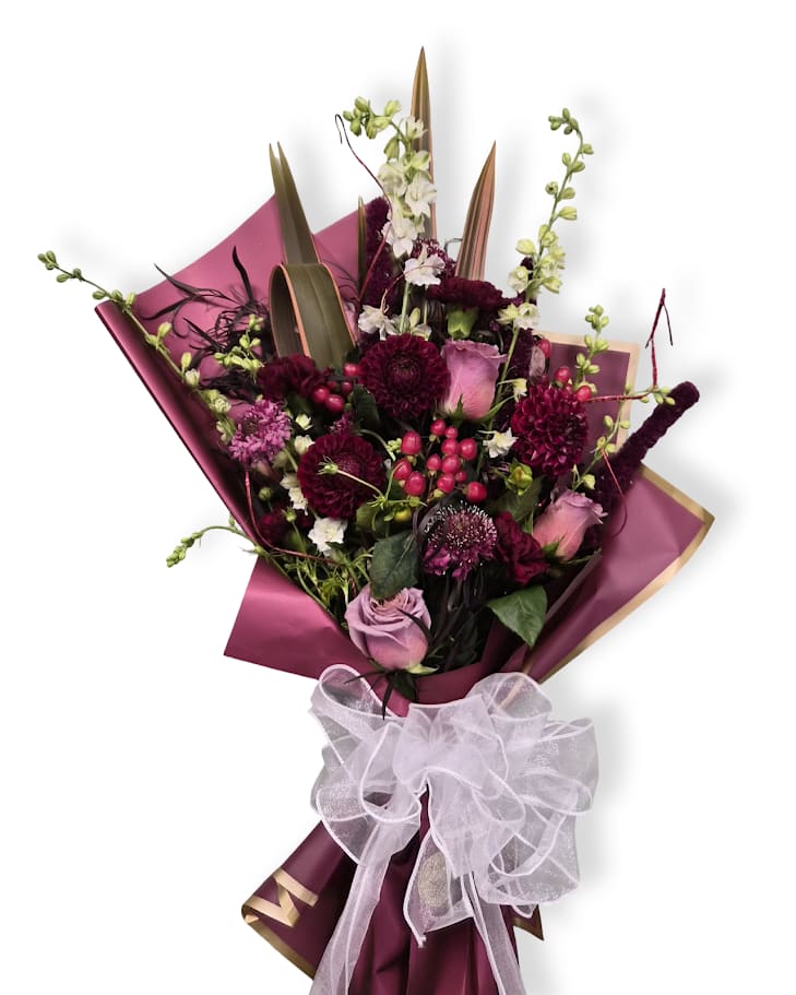 Dark Ruby Cello Wrap - Deep burgundy dahlias and roses mingle with soft pink roses and lavender-toned blooms for a rich, romantic color story-the deep ruby, playful lavender, crisp white, and fresh green create a striking, celebratory palette perfect for an anniversary, birthday (ruby-month), or a thoughtful just-because gift.  This hand-tied arrangement features roses, dahlias and complementary seasonal stems arranged with a dense, layered center and taller pale green and white larkspur for a garden-style silhouette. Wrapped in magenta decorative paper and finished with a sheer organza ribbon, it arrives polished and ready to gift.  Choose this bouquet when you want her to feel noticed-bold like a ruby, whimsical like larkspur. Simple care: trim stems and keep fresh water in a vase after unwrapping to help blooms last. Vase not included.