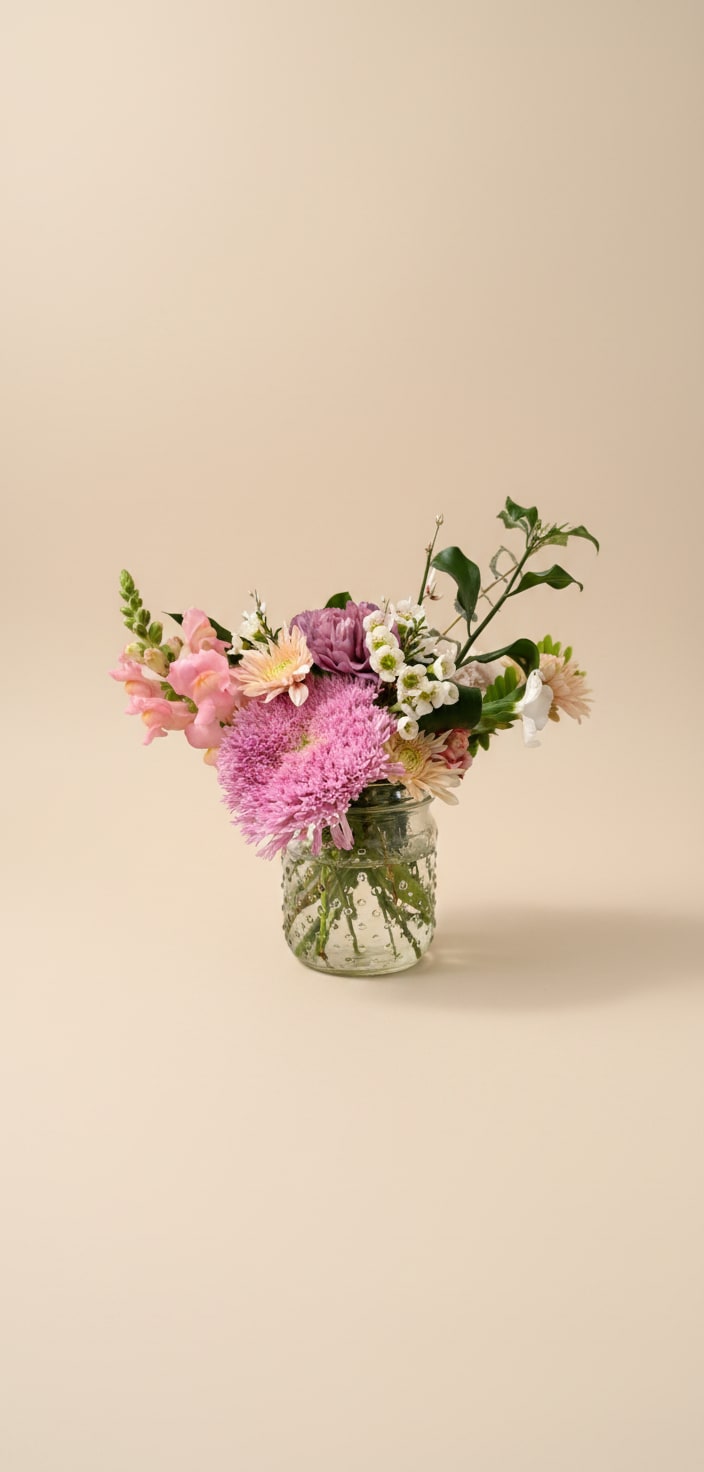 Bloom Jar - Soft pink and white blooms gathered in a clear glass mason jar give this petite flower arrangement a calm, loving presence-perfect little way to say I love you. The Bloom Jar reads like a garden-picked moment: the recipient will feel gently celebrated by its soft palette and warm peach accents that bring a quiet, affectionate energy to any room.  Rounded chrysanthemum-like blooms sit alongside delicate white filler flowers and variegated green foliage for a layered, textured look that feels both full and airy. Arranged in a simple clear glass jar, the composition highlights the stems and keeps the focus on the flowers' soft pinks, creams, and subtle peach tones. Sized for tabletops, desks, or bedside tables, this everyday vase arrangement needs only fresh water and a bright spot out of direct afternoon sun to stay lovely for days-an easy, elegant way to make someone feel truly seen.  Every effort will be made to recreate your chosen design. Please note that from time to time due to product availability and quality we may need to make substitutions. We will make sure to keep the original design in mind when making substitutions.
