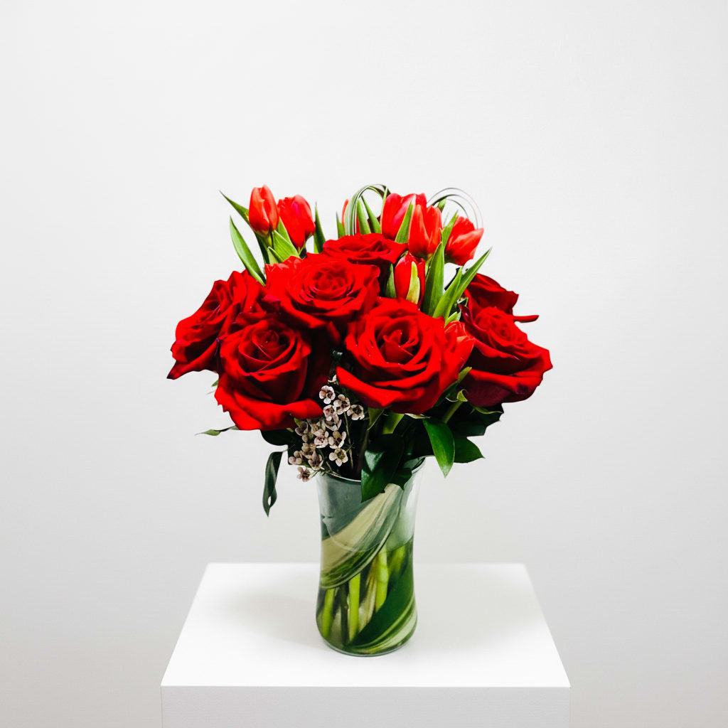 True Romance Arrangement Red Roses and Tulips  - A dense cluster of velvety red roses and red‑orange tulips creates a bold, romantic statement perfect for an anniversary bouquet or any love and romance moment. The tall clear glass vase gives the arrangement contemporary height while broad green leaves wrapped inside the vase conceal stems and add a structured, modern touch; small white filler blooms peek through the red for delicate contrast and texture.  This arrangement blends deep reds, warm orange accents, crisp white highlights and fresh greenery for a rich, monochromatic look that reads dramatic and refined. Sized for impact in a tall vase, it makes a meaningful birthday surprise, a thinking‑of‑you gesture, or a celebratory gift for congratulations or Valentine's Day - the person you send it to will feel genuinely celebrated and seen. Care: keep water fresh and display away from direct heat to prolong bloom life.