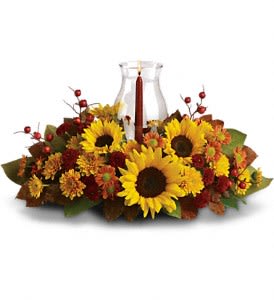 Sunflower Centerpiece - Symbolizing warmth and happiness, it is no wonder the sunflower is a quintessential ingredient in so many fall bouquets. This centerpiece maximizes the joy of sunflowers by arranging them with a bevy of fall flowers, foliage and a cranberry hued candle under hurricane glass.  Along with brilliant sunflowers, you'll find yellow spray roses, bronze and rust chrysanthemums, red berries, magnolia leaves and of course an elegant tapered candle inside a hurricane vase.  Approximately 21" W x 12" H  Orientation: All-Around