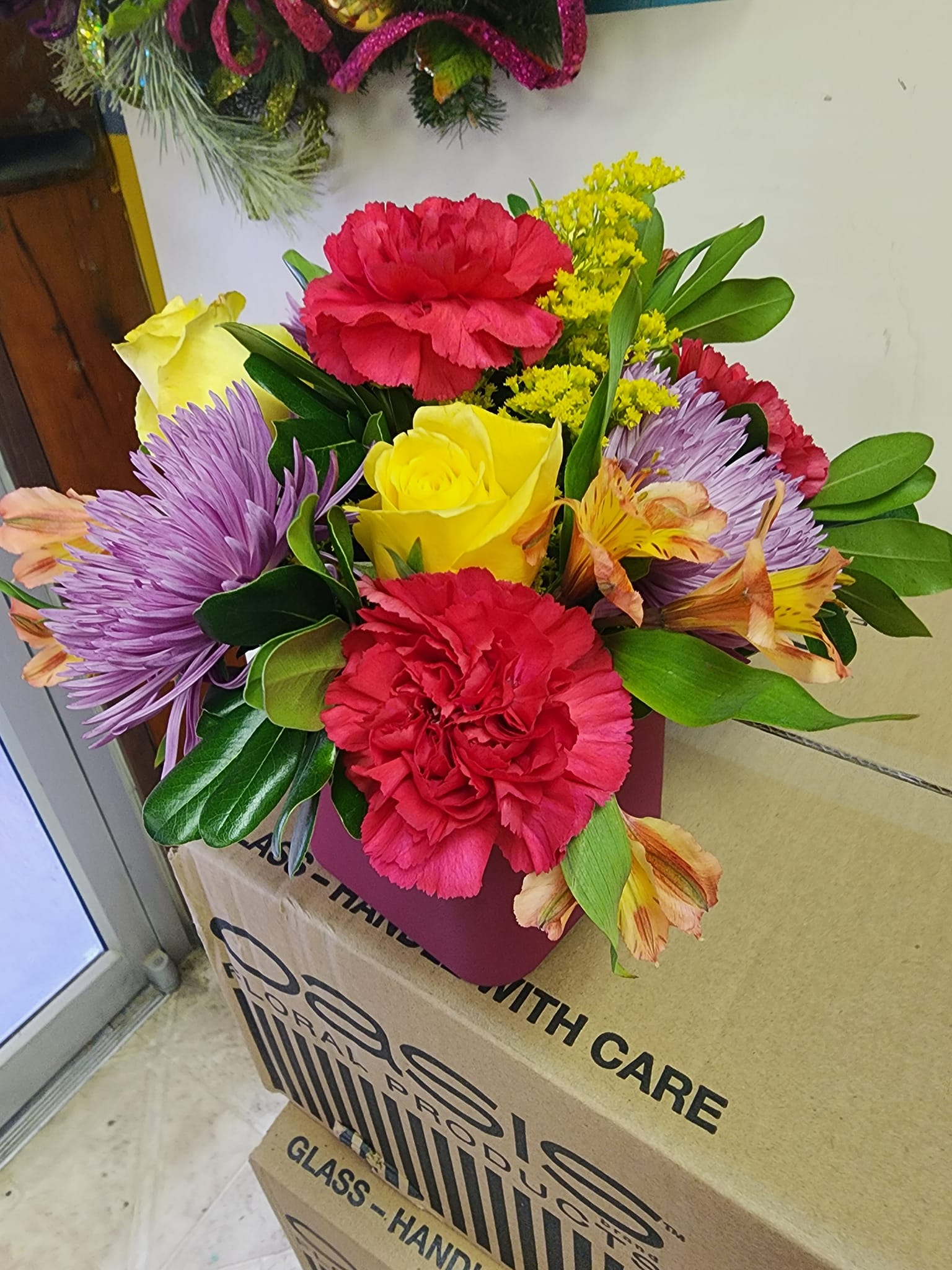 Mixed bouquet with red carnations, yellow roses, and purple flowers in a pink box