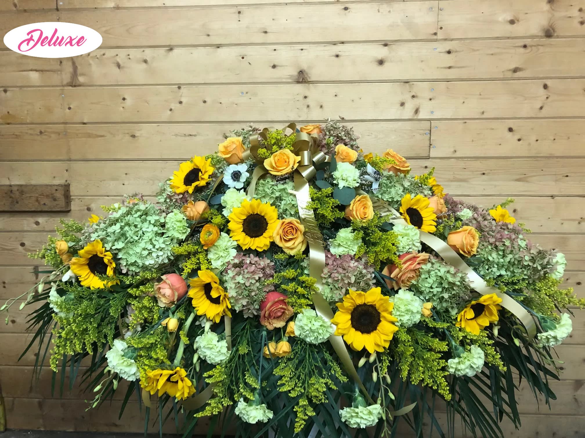 Sunny Isle Casket Cover  - Sunny Isle is a beautiful casket cover designed to honor a cherished life with warmth, love, and joyful remembrance. This vibrant floral tribute features radiant sunflowers, striking orange roses, lush green hydrangea, soft green carnations, and cheerful yellow accent flowers, thoughtfully arranged to create a natural expression of peace, hope, and gratitude. Inspired by the beauty of a sunlit garden, the rich blend of golden yellows, vibrant oranges, and fresh greens celebrates a life filled with happiness, kindness, and treasured memories. Handcrafted by our compassionate florists, Sunny Isle offers a heartfelt way to celebrate a loved one’s unique spirit while providing comfort and support to family and friends during a time of loss.
