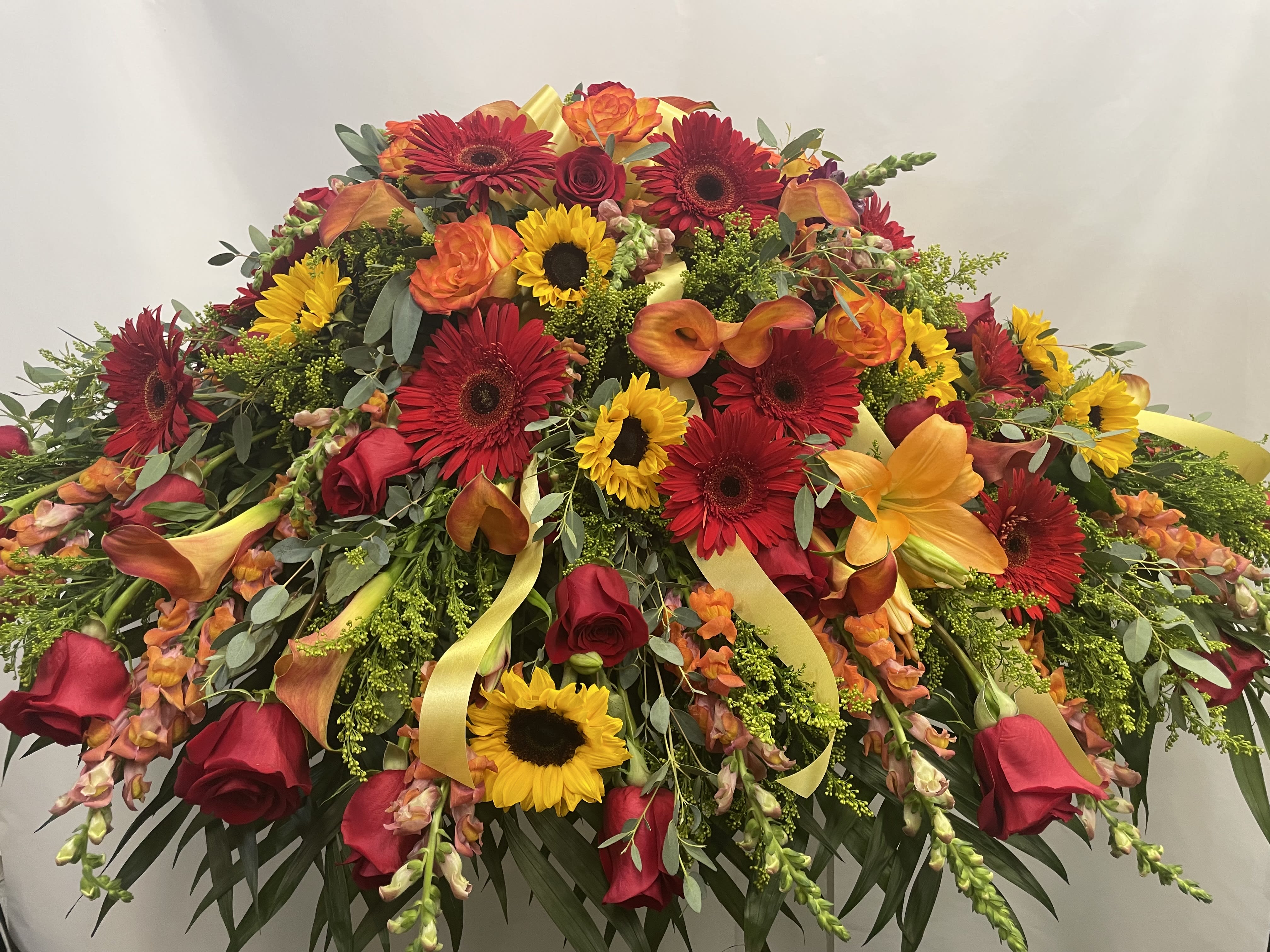 Autumn's Embrace  - Celebrate a life filled with warmth, love, and cherished memories with Autumn's Embrace, a radiant casket cover inspired by the rich beauty of the changing seasons. This striking tribute features vibrant orange lilies, orange snapdragons, brilliant red gerbera daisies, classic red and orange roses, cheerful sunflowers, delicate goldenrod, and fragrant eucalyptus, creating a stunning display of warmth, color, and heartfelt remembrance.  Symbolizing admiration, gratitude, and enduring love, this thoughtfully designed arrangement offers a comforting expression of honor for a life beautifully lived. Handcrafted by our skilled florists using the freshest flowers available, Autumn's Embrace serves as a meaningful final tribute, celebrating a legacy of love with grace, beauty, and lasting remembrance. 