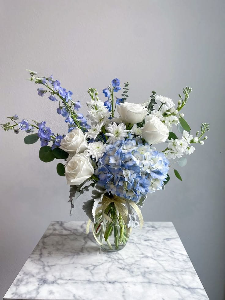 Tiny Treasure - Tiny Treasure is a graceful fresh flower arrangement created to celebrate the arrival of a precious baby boy. Featuring lush blue hydrangea, classic white roses, and delicate light blue delphinium, this handcrafted design is artfully arranged in a tall clear glass vase for a timeless and elegant presentation. The soft shades of blue and white create a peaceful, welcoming display that makes a thoughtful gift for new parents, baby showers, or welcoming a little one home. Whether sent to a hospital or enjoyed in the nursery, Tiny Treasure is a beautiful floral gift that expresses love, joy, and heartfelt congratulations while creating a lasting impression.