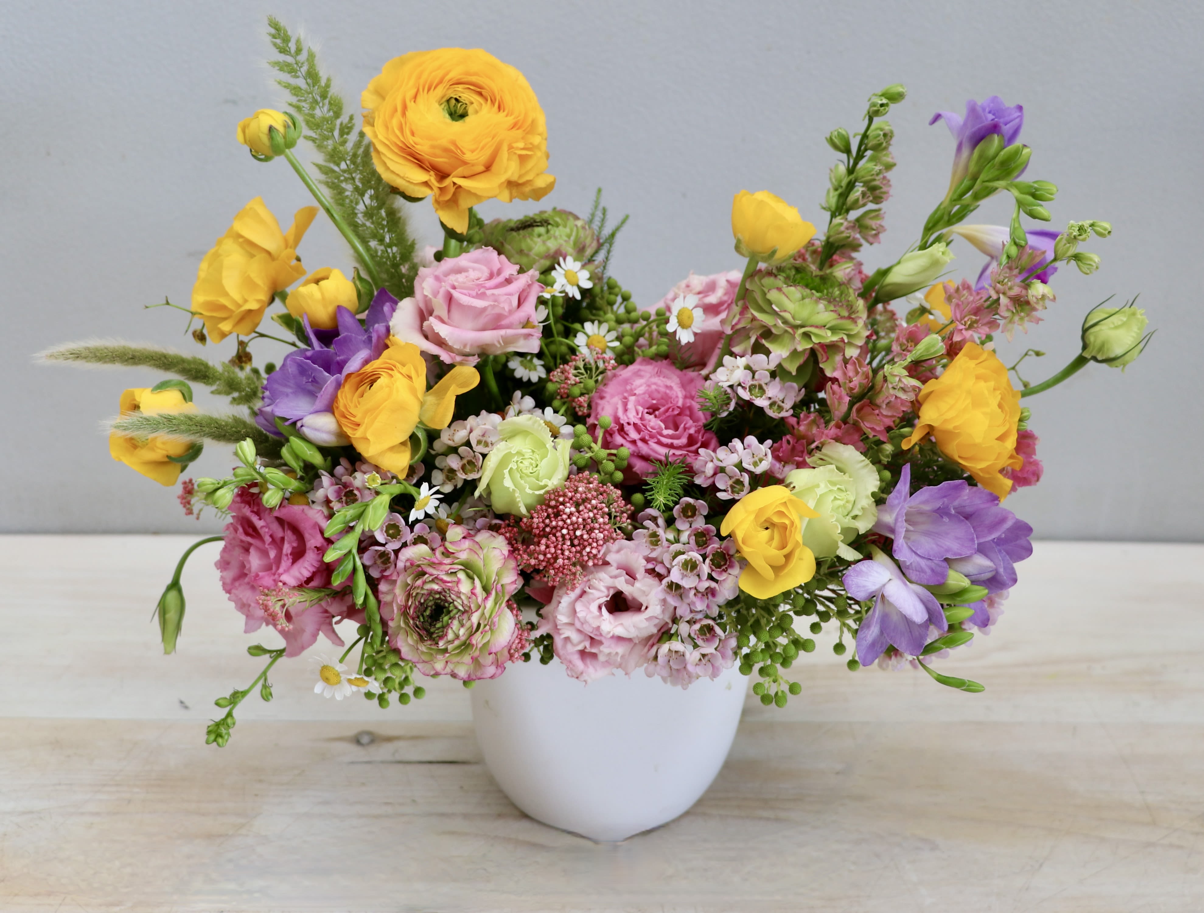 Low arrangement of yellow ranunculus, pink blooms, and purple flowers in a white bowl vase