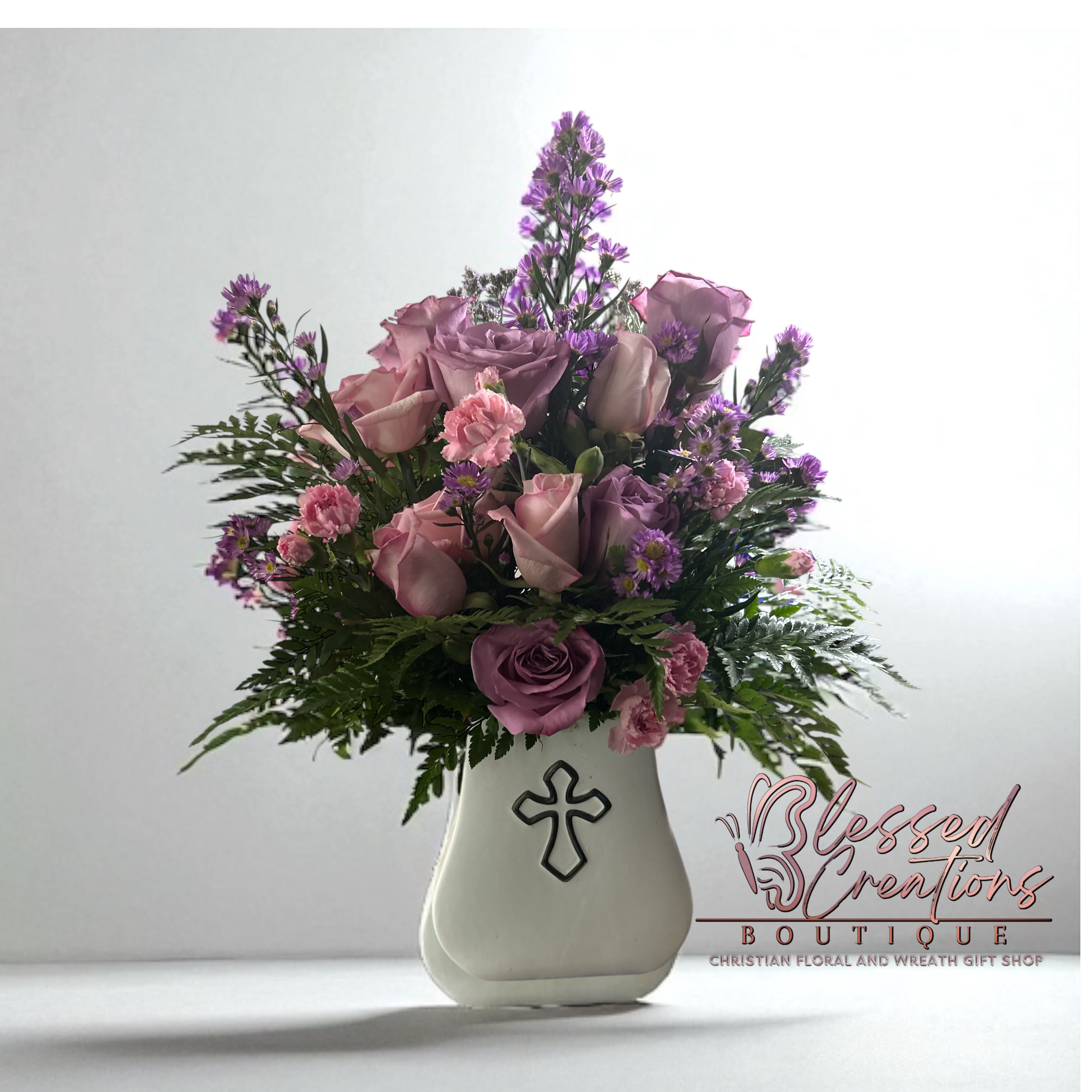 Pink and lavender roses with purple filler flowers in a white vase with a cross