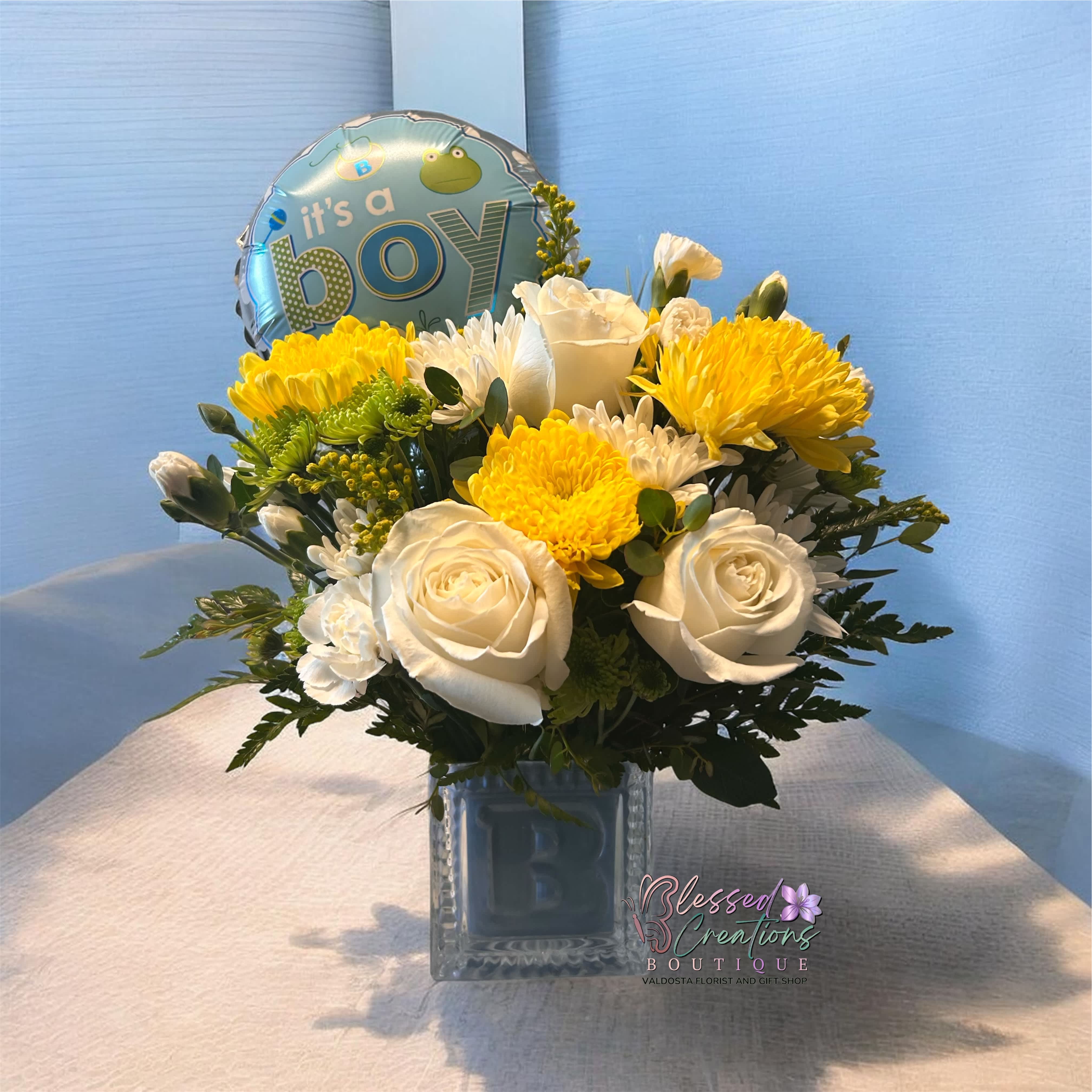 White and yellow flower arrangement in a glass cube vase with a boy balloon