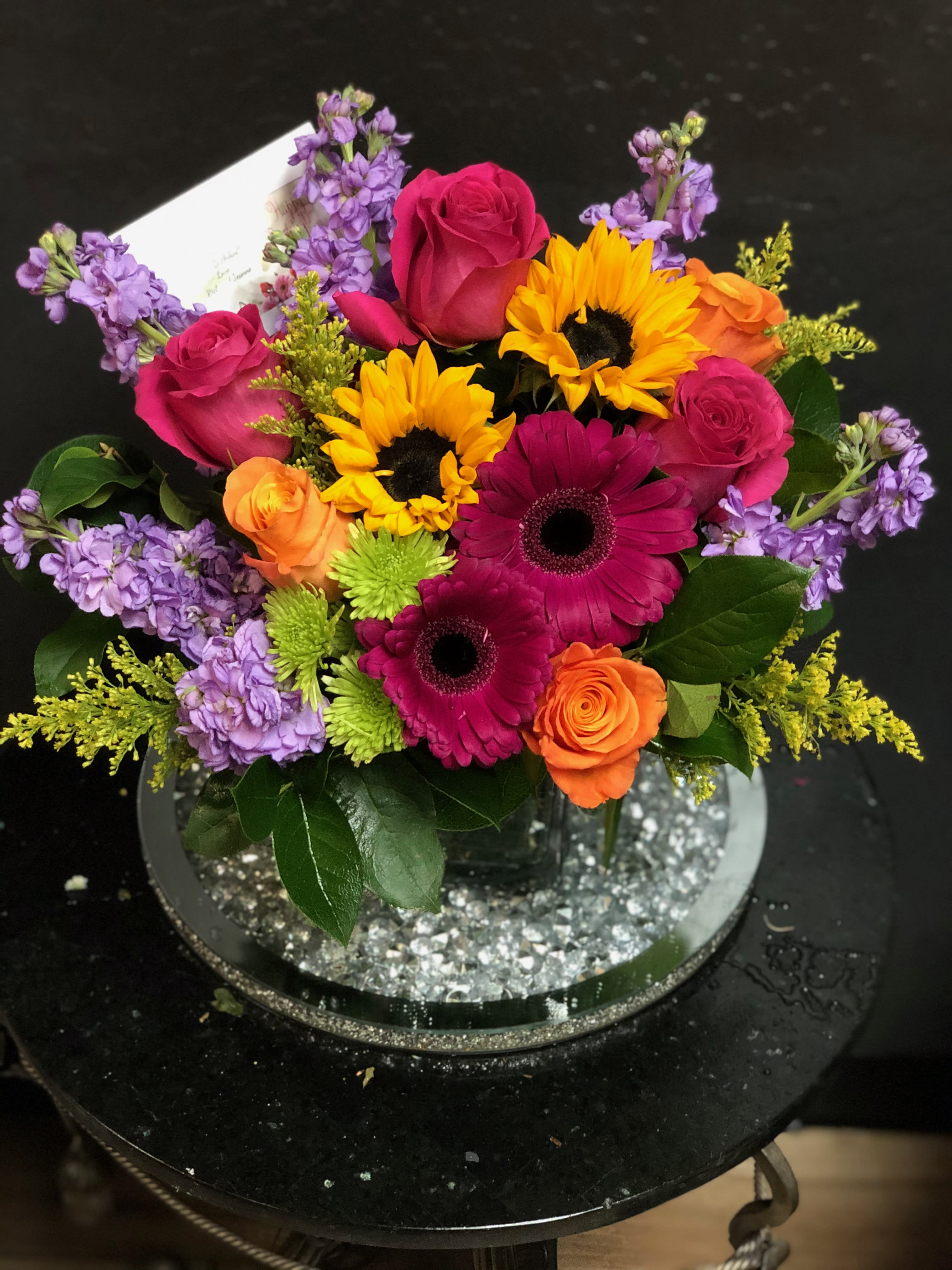 Bright mixed bouquet with roses, gerbera daisies, sunflowers, and purple blooms in a vase