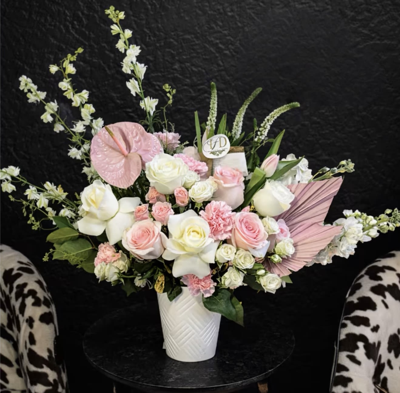 Empress Pink Bouquet  - Soft blush roses and sculptural anthurium leaf make a memorable first impression in this flower arrangement designed for birthdays and welcoming a new baby. The Empress Pink Bouquet reads as a polished, gift-ready statement-its romantic mix of pink, white and green blooms plus a decorative pink folded fan and round logo pick will make the recipient feel truly celebrated.  This mixed bouquet gathers large roses and tulips with carnations, calla lilies and clustered filler flowers for a lush, textured look. Tall white accent stems add graceful movement while the compact, textured white ceramic vase keeps the design neat on a bedside table or dining room centerpiece. Colors range from pale blush to deeper pinks against fresh greenery for a versatile, season-spanning palette.  Perfect for congratulating new parents, honoring Mom on Mother's Day or sending a thoughtful birthday message, this arrangement arrives ready to display - no extra vase required. Place in a cool spot away from direct sun and refresh vase water every two days to help blooms last longer.
