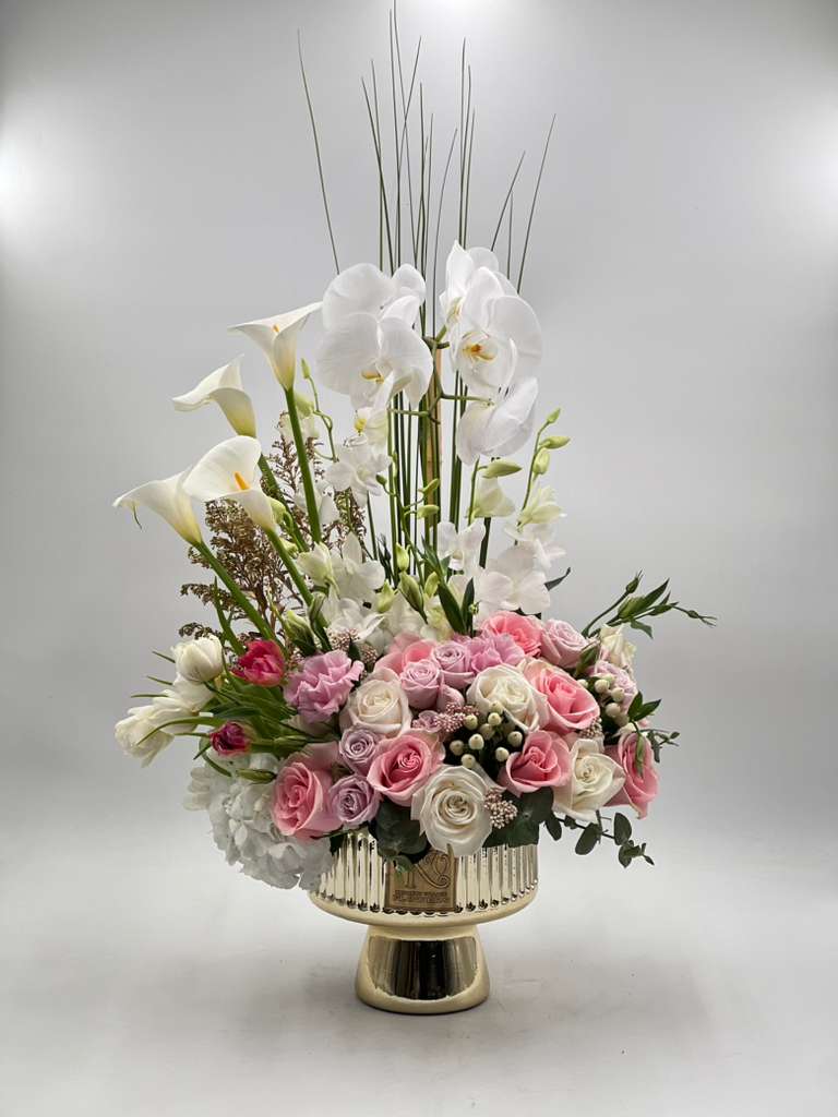 Grand Elegance Arrangement - A striking statement of elegance and sophistication, this luxurious floral arrangement features graceful white orchids and calla lilies rising above a romantic garden of blush, pink, ivory, and lavender roses, accented with delicate seasonal blooms and lush greenery. Designed in a reflective pedestal vase, it brings height, softness, and modern luxury together in one unforgettable piece. Perfect for anniversaries, grand celebrations, sympathy expressions, birthdays, or upscale gifting, this arrangement is created to leave a lasting impression with its refined beauty and graceful presence.