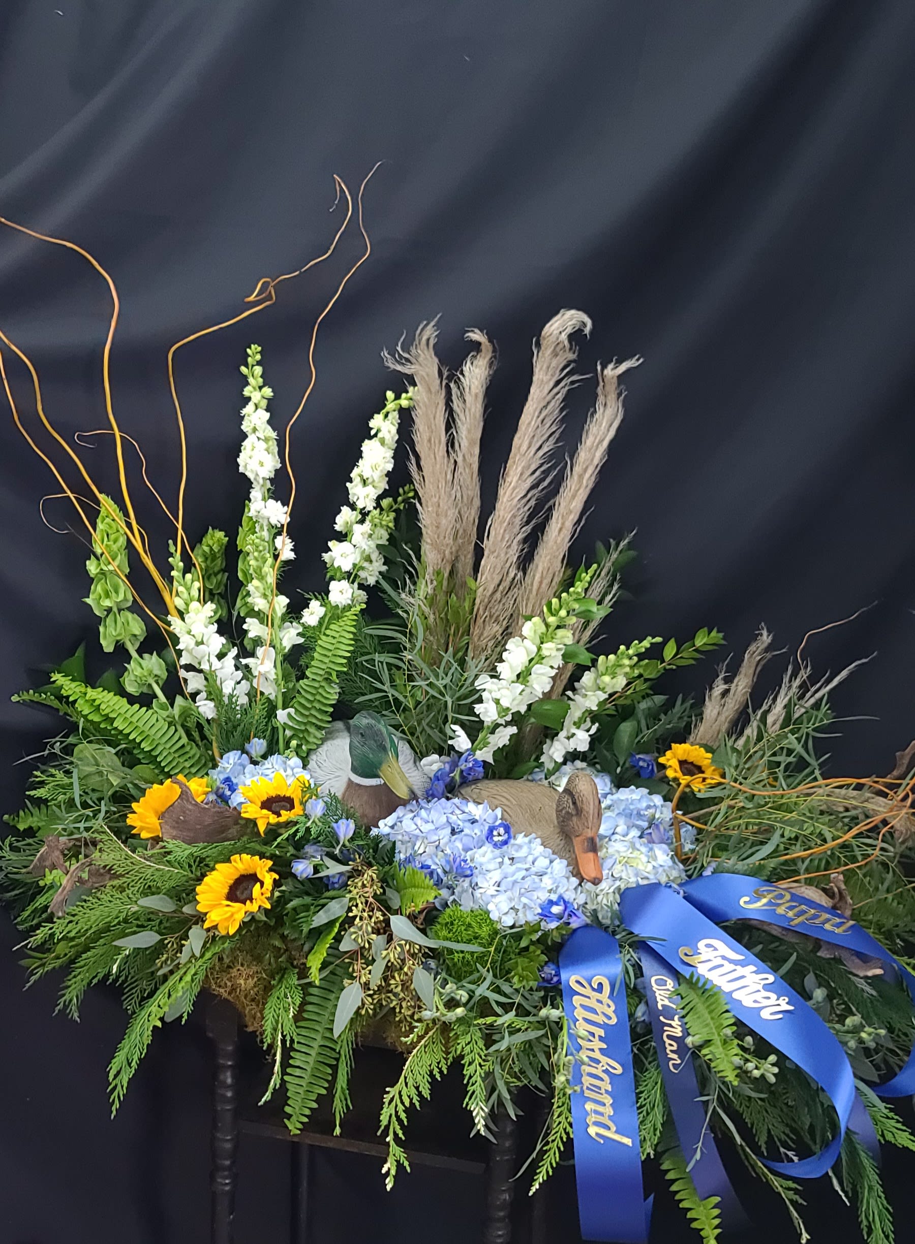 Floral tribute with sunflowers, blue hydrangeas, and blue ribbons on a dark stand