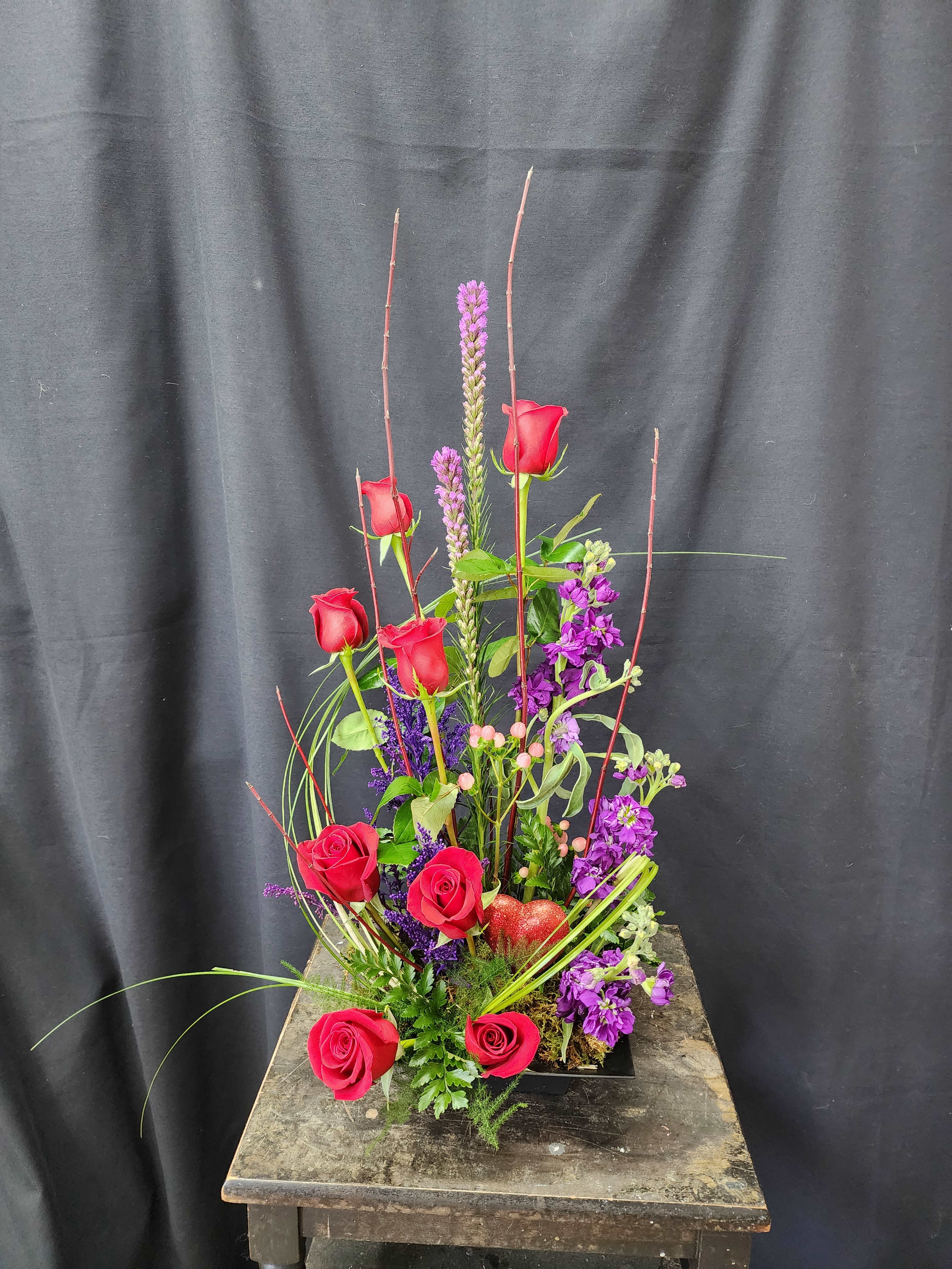 Tall arrangement of red roses and purple flowers in a low black container
