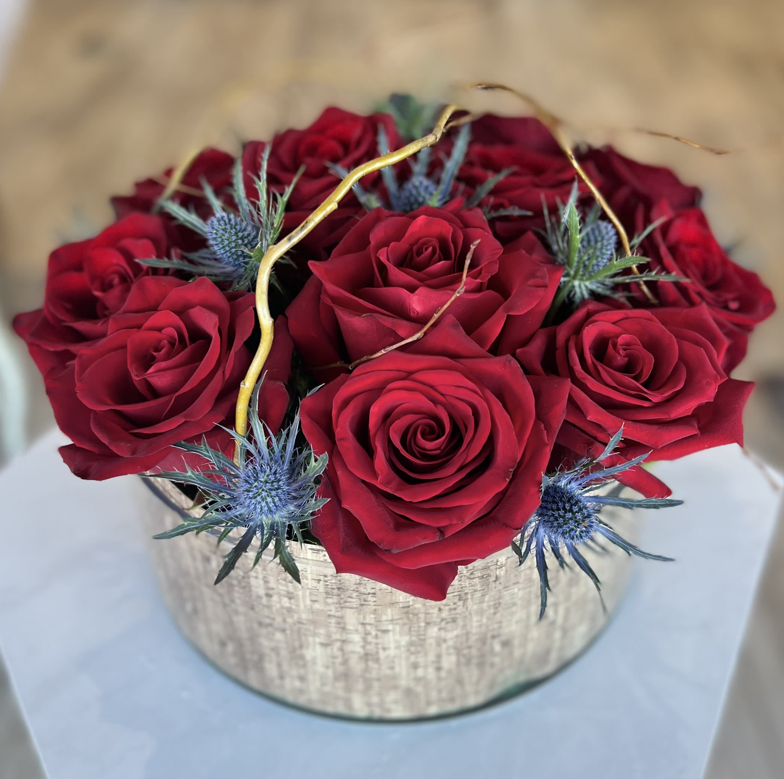 Red roses arranged in a round silver box with blue thistle accents
