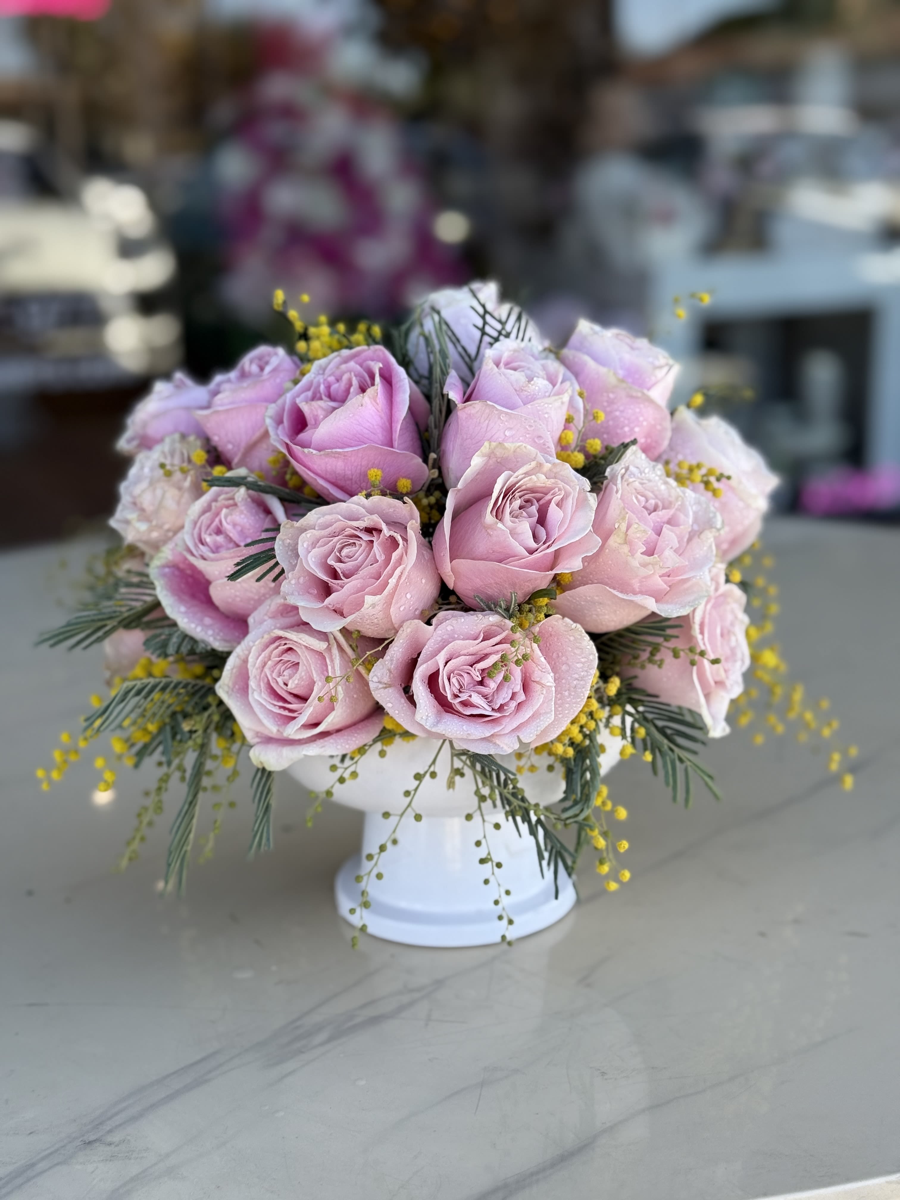 Pink roses arranged in a white pedestal vase with yellow filler flowers