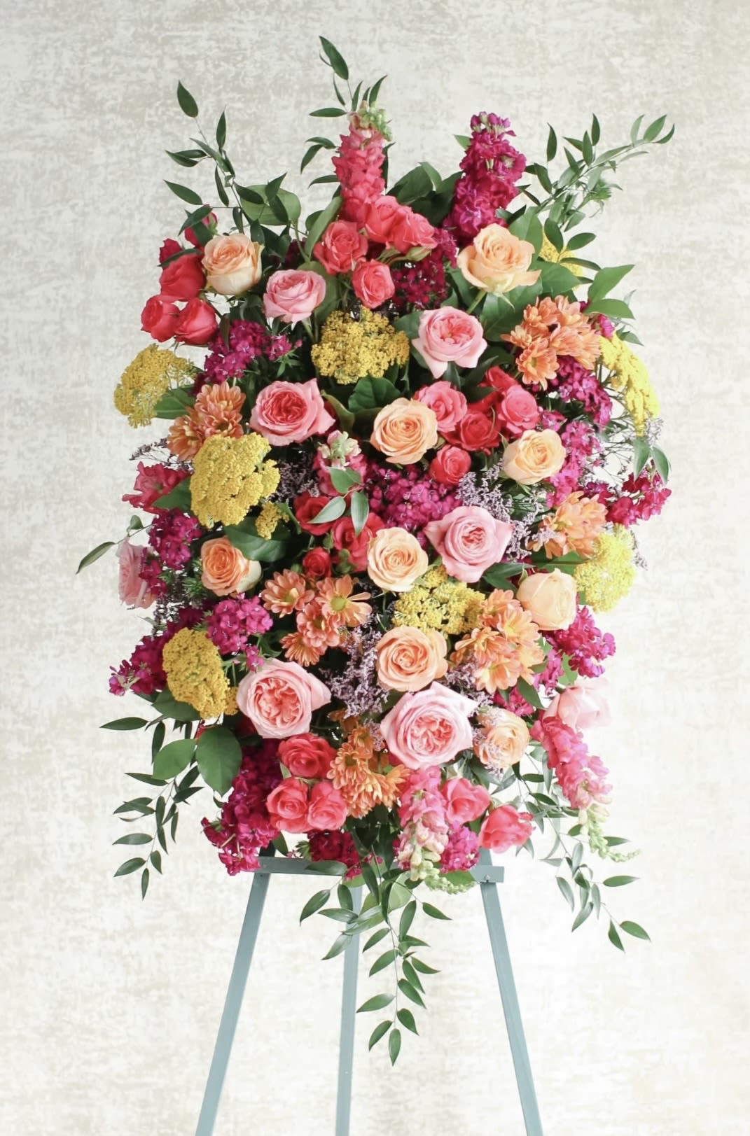 Colorful Funeral Spray - A large upright standing spray that reads as both a heartfelt tribute and a celebration of a life well-lived - this funeral spray brings together roses, hydrangeas, chrysanthemums and snapdragons in mixed colorways of pink, peach, red and bright yellow. Designed for sympathy and funeral services, the bold vertical spikes and dense, layered composition create a dramatic silhouette on an easel stand that commands attention at the front of a service or beside a casket.  The arrangement is packed with garden-style roses in warm pinks, peaches and reds, with clusters of hydrangea for volume, cheerful yellow and magenta filler blooms, and textured chrysanthemums and snapdragons to add height and movement. Trailing greenery softens the edges and gives the spray a full, finished look; the dense, layered design favors visibility from a distance and photographs beautifully in a memorial setting.  Ideal for honoring someone who loved vibrant color or whose life you want remembered with warmth and joy, this spray expresses sympathy while celebrating personality - a fitting choice for funerals, memorial services, or a bright floral tribute at a graveside. It arrives ready on an easel stand; for delivery and placement details or to request personalization (ribbon colors or a message card), please contact the shop.