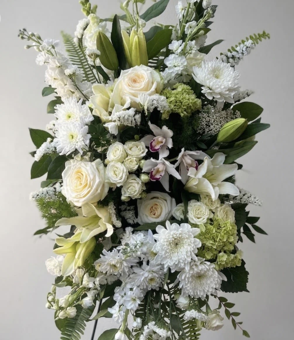 Elegant Funeral Spray  - A tall, layered standing spray that brings quiet dignity to a funeral or memorial service, this sympathy arrangement conveys calm and respect with its white, green, and purple palette. The formal, symmetrical composition of cream roses and white lilies reads as a classic funeral spray, while the soft green clusters and airy branching elements add lift and a living, peaceful texture-an arrangement meant to comfort the person you're honoring.  Cream roses, white lilies, and white chrysanthemums form the heart of the design, accented by green, hydrangea-like blooms and delicate orchids with purple centers for a subtle touch of contrast. The spray is mounted on a standing easel and arranged in tiers to present clean lines and an uplifting silhouette suitable for a chapel or graveside display. Its serene colors and layered form make it a respectful choice for expressing sympathy and remembrance.
