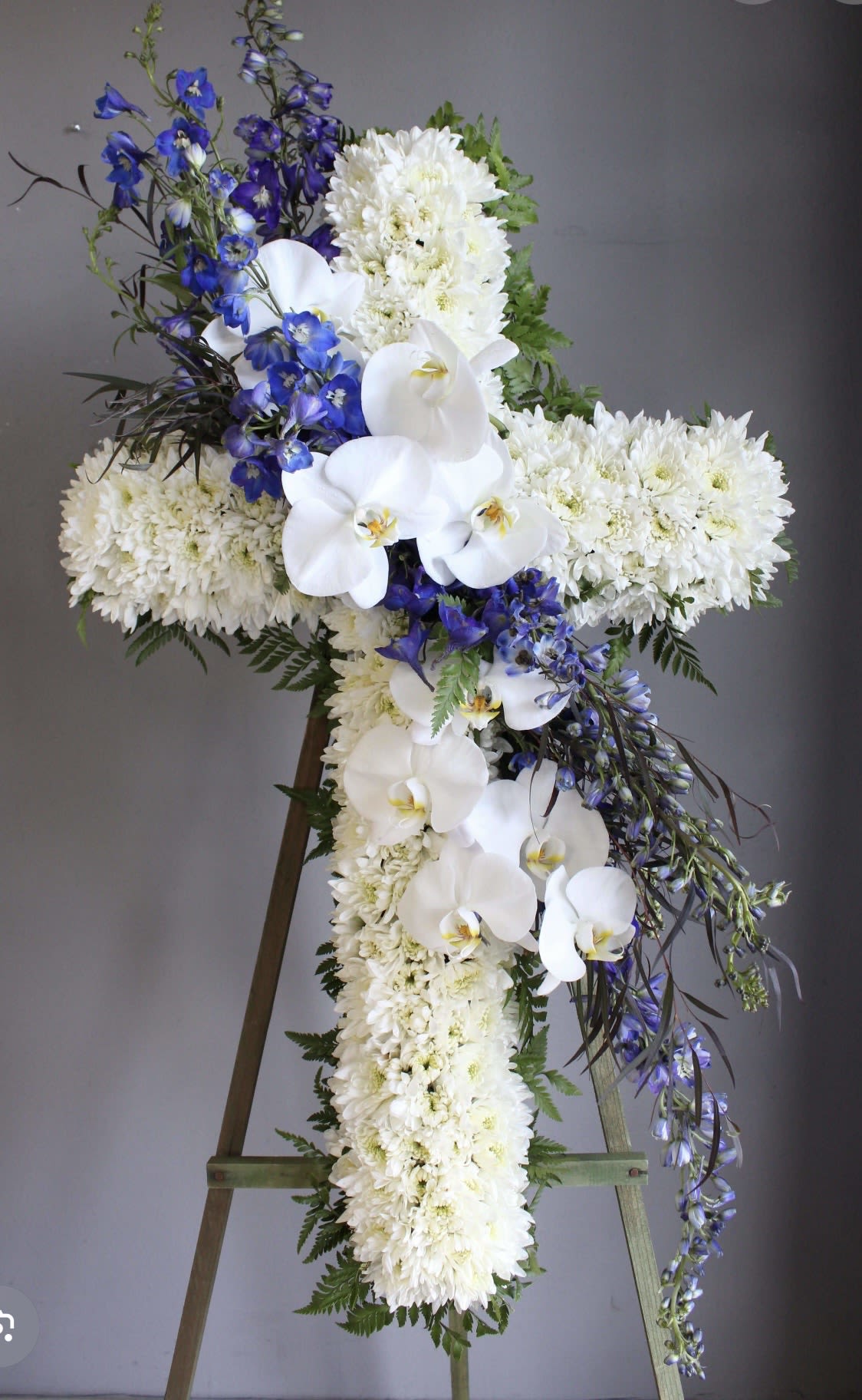 Funeral Cross white and blue - A dignified, devotional cross crafted for a memorial or funeral service - this flower arrangement is a white and blue standing cross designed to convey reverence and sympathy. The cross shape made of dense white chrysanthemums provides a serene backdrop while white Phalaenopsis orchids are layered across the center and lower section to create a gentle focal point. Bright blue delphinium sprays rise through the design, giving vertical movement and a calm contrast that reads peaceful and respectful.  Made to display on a wooden easel, the arrangement reads formal from a distance and detailed up close: the chrysanthemums form the solid silhouette, orchids add luxurious texture, delphinium bring cool blue accents, and touches of greenery soften the edges. Its tall presentation and classic color palette make it appropriate for a funeral service, memorial gathering, or graveside tribute - a meaningful way to honor the person you're remembering.  If you need help with delivery timing for a service or placement instructions for the easel, we'll coordinate details so the tribute is presented exactly when and where it's needed. Care is minimal once displayed; the standing cross is arranged to remain dignified throughout the service.