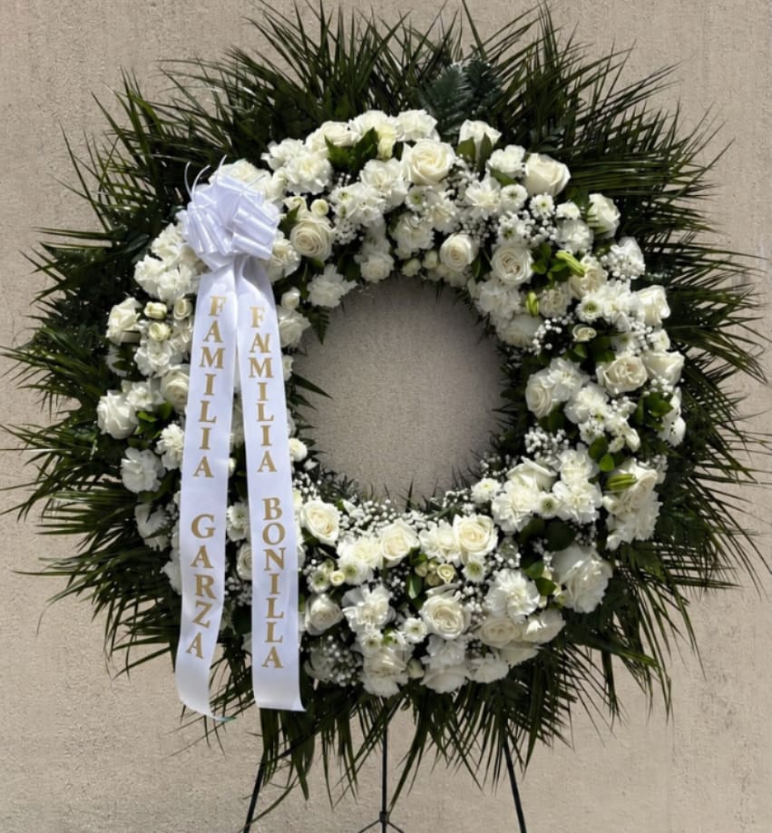 LARGE Wreath - A large, formal sympathy wreath designed to offer a solemn, respectful tribute at a funeral or memorial service. This standing wreath arrangement features mostly white blooms-roses, carnations, chrysanthemums and small daisy-like flowers-set against a dramatic halo of dense, dark green, palm-like foliage. The white ribbon streamers with gold lettering add a personalized memorial touch that underscores the wreath's purpose as a meaningful remembrance piece.  Generous and traditionally styled, the wreath is built on a standing wreath frame and is shown displayed on a metal easel stand, making it suitable for church services, viewings, or graveside presentations. The dense, circular composition and contrasting textures-the soft, round flowers against spiky green foliage-create a serene visual impact that helps the person you're honoring feel revered and remembered. Available in multiple sizes (small, medium, large) and finished with white ribbon streamers; ordering includes the standing wreath frame and metal easel as shown.