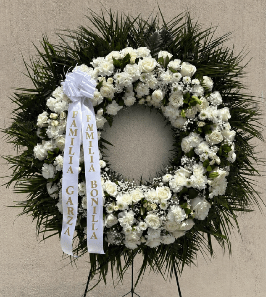 SMALL WREATH - A classic white sympathy wreath that offers a quiet, dignified tribute for a funeral or memorial service. This circular funeral wreath features white roses, carnations, chrysanthemums and daisy-like blooms arranged in a densely packed ring so the floral surface reads as a soft, luminous cloud; dark green palm-style foliage frames the outer edge to create striking contrast and a formal silhouette the family will notice.  Available in small, medium and large standing sizes, each wreath comes mounted on a metal easel stand with a floral wreath stand and finished with white ribbon streamers-some examples include gold-lettered memorial ribbons as shown. The texture of ruffled carnations and the creamy petals of roses combine for a gentle fragrance and layered look that reads respectfully in photos or at a graveside.  Made to express sympathy and honor, this wreath is the kind of arrangement that helps the person you're remembering feel held and recognized. Order with delivery to a funeral home or cemetery; if you need a custom ribbon inscription or specific size, please indicate that when placing your order.