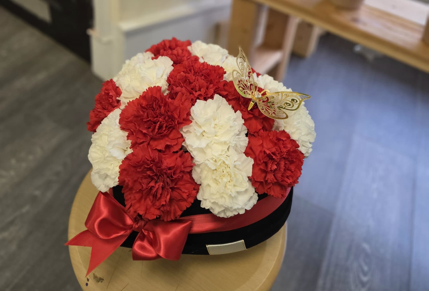 Red and white carnation arrangement in a black hat box with a red ribbon and butterfly decoration