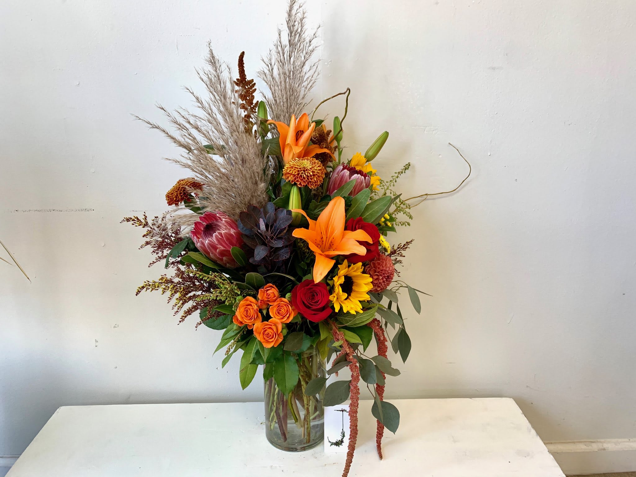 Tall mixed bouquet with orange lilies, red roses, and sunflowers in a glass vase