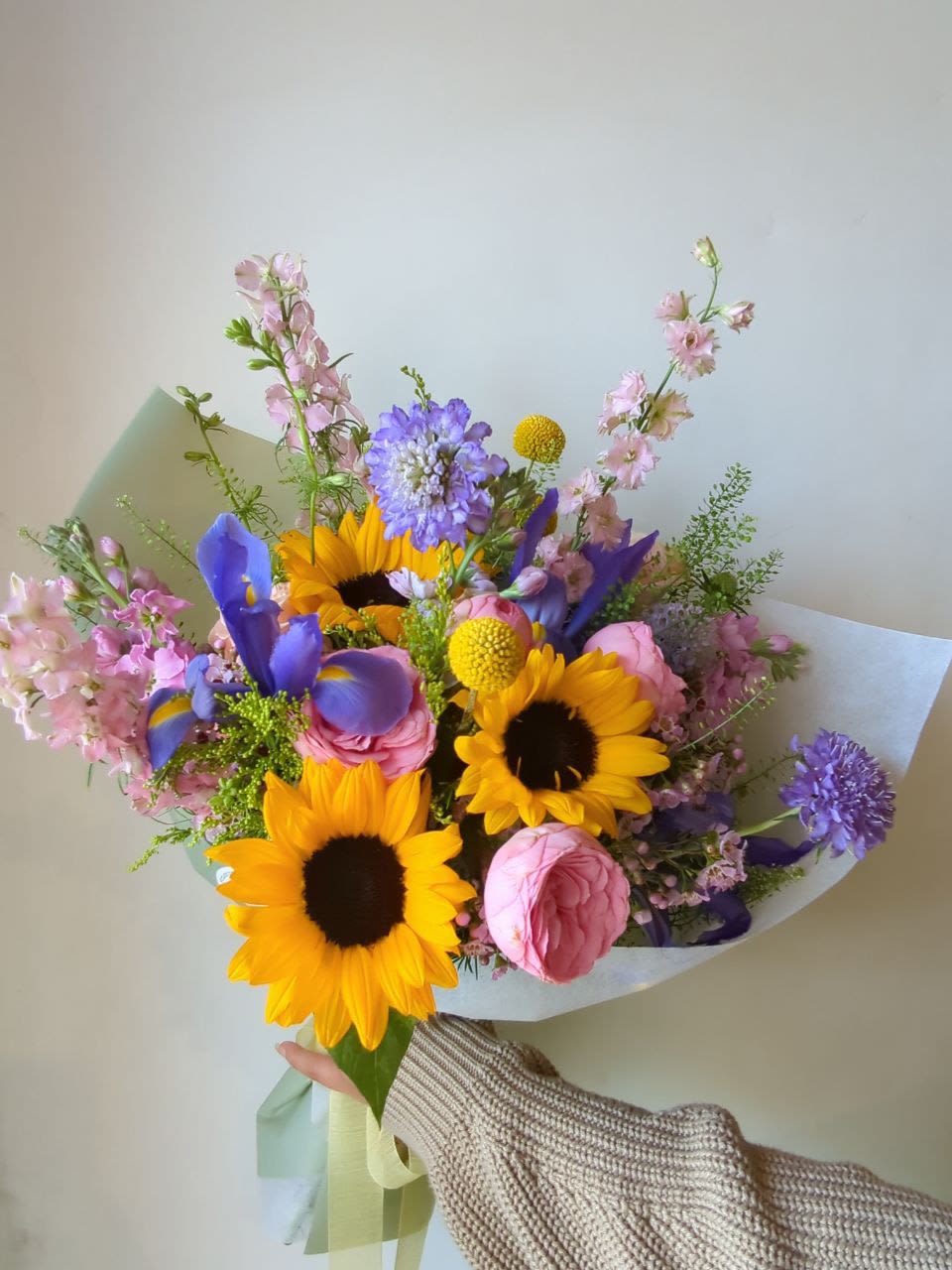 Handheld bouquet with sunflowers, pink roses, and purple blooms wrapped in paper