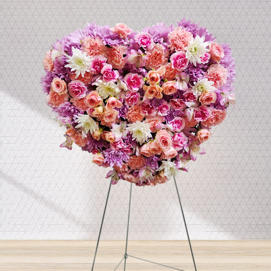 In Loving Radiance by Latin Lady Flowers - In Loving Radiance is a stunning heart-shaped funeral spray crafted to express deep love and tender remembrance. This elegant arrangement features a harmonious blend of peach carnations and roses, complemented by lavender chrysanthemums and daisies, soft pink carnations, pristine white chrysanthemums, and delicate pink alstroemeria. The gentle pastel palette symbolizes grace and serenity, offering a heartfelt tribute to a cherished life. Perfectly designed to convey compassion and solace, this floral masterpiece is a touching way to honor and celebrate the memory of a loved one.
