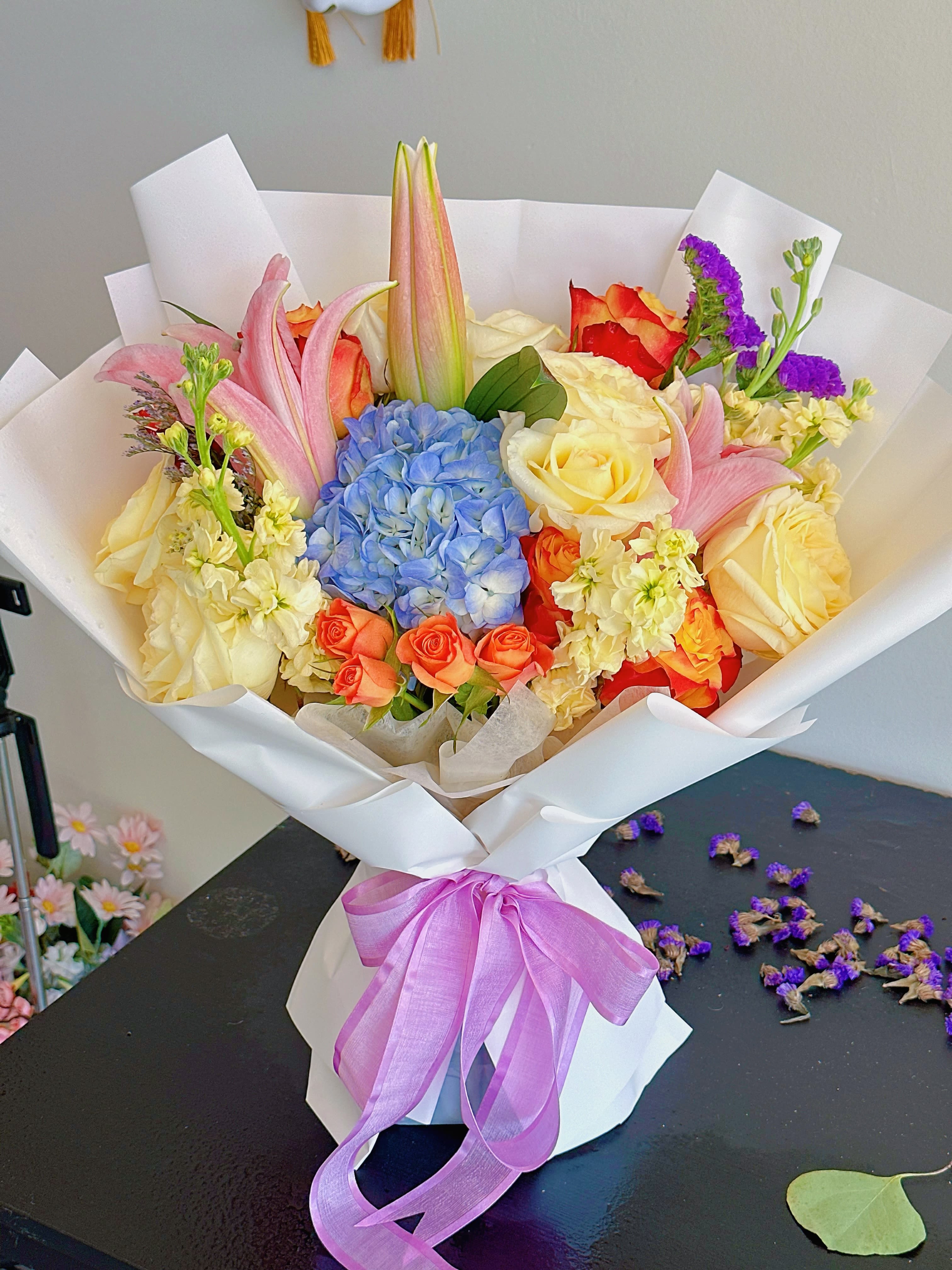 Bouquet of roses, lilies, and blue hydrangea wrapped in white paper with a purple ribbon